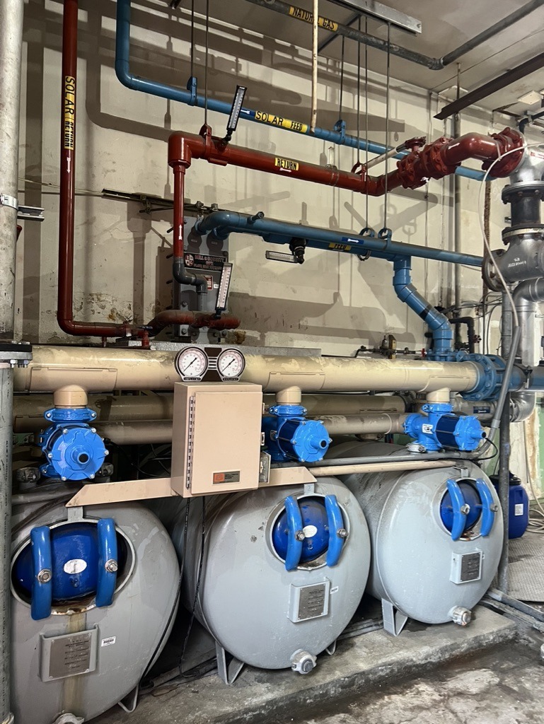 Industrial room with complex pipes, gauges, and valves. Blue and red pipes run horizontally and vertically, connecting machinery and a control box.