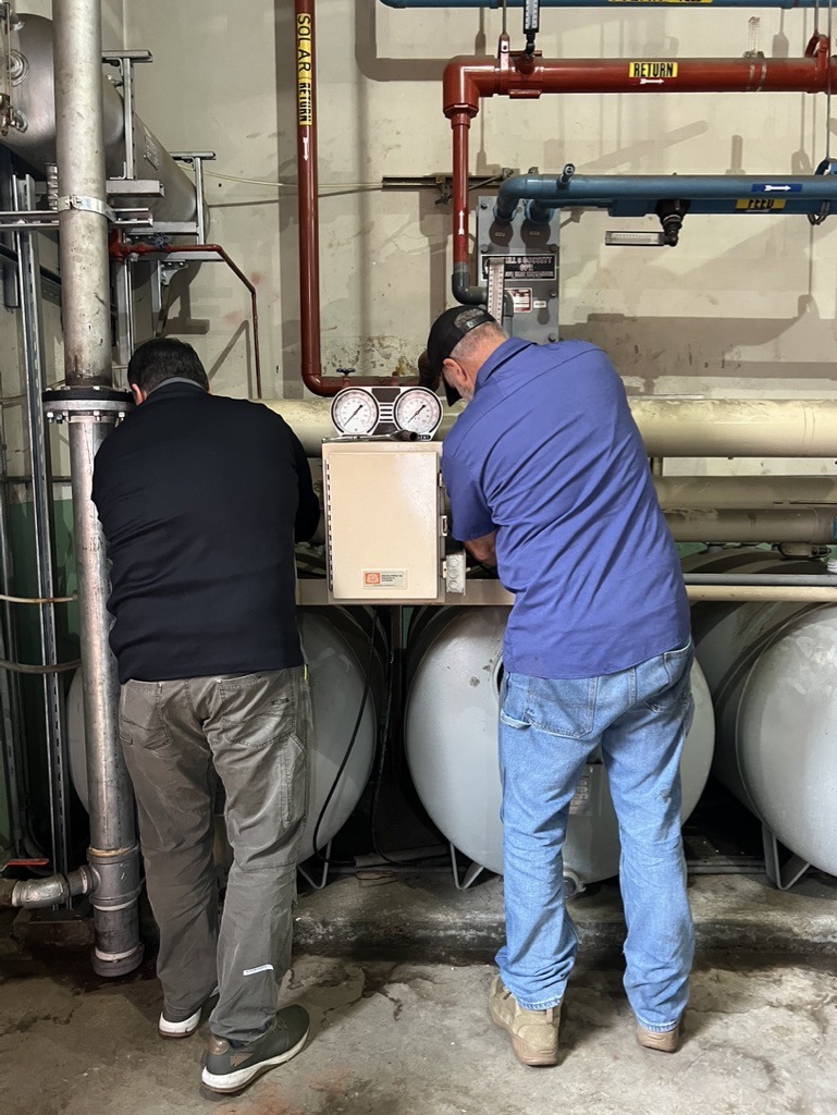 Two people are working on industrial piping with gauges in a utility room. They appear to be engaged in maintenance or repair tasks.