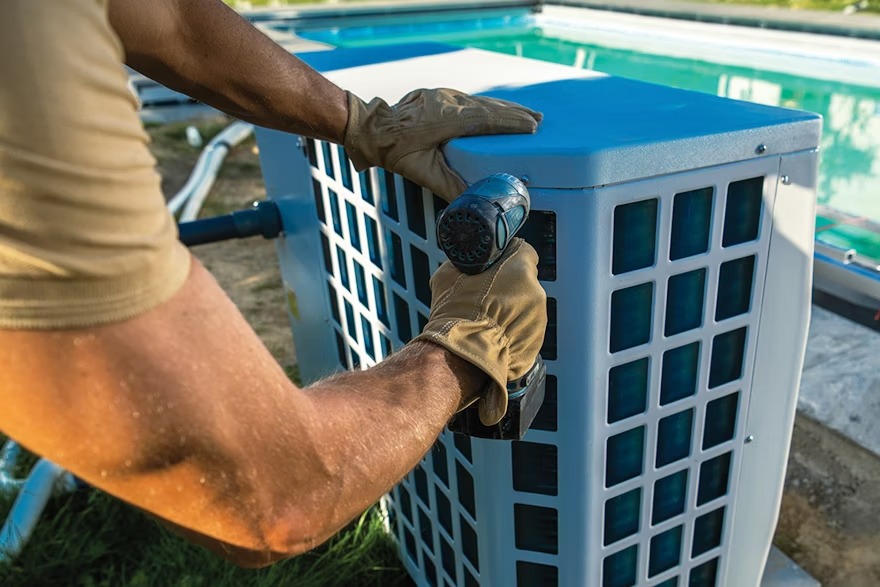 A person wearing gloves uses a drill on a poolside outdoor air conditioning unit, surrounded by greenery and a clear swimming pool.