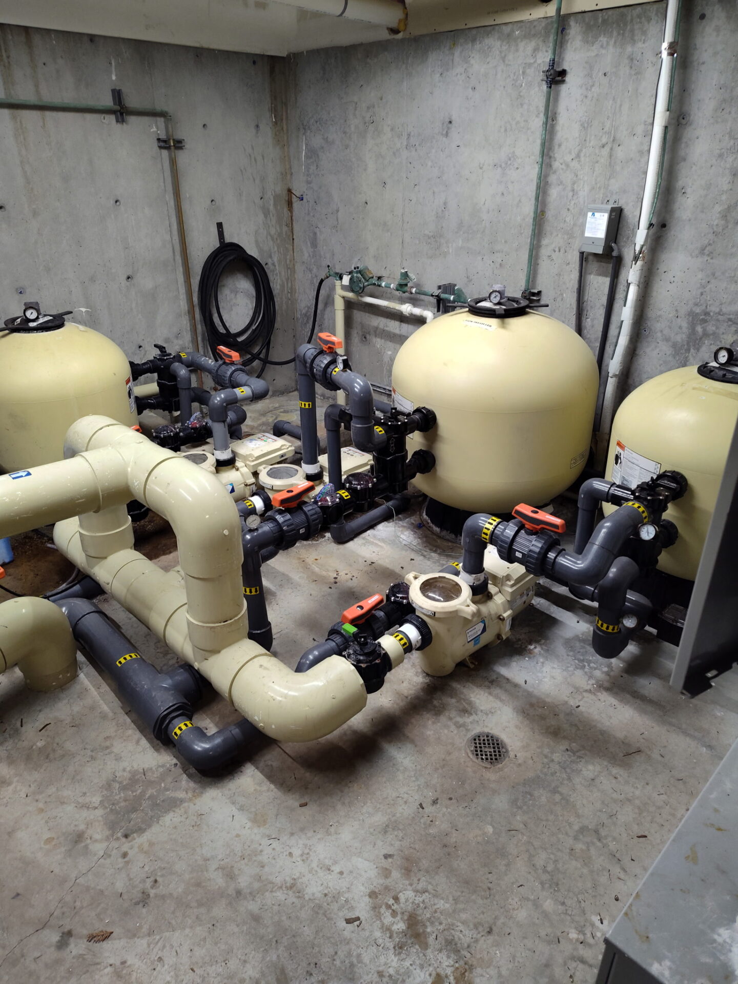 Room with large beige filtration tanks and numerous interconnected pipes, valves, and controls, likely part of a water or pool processing system.