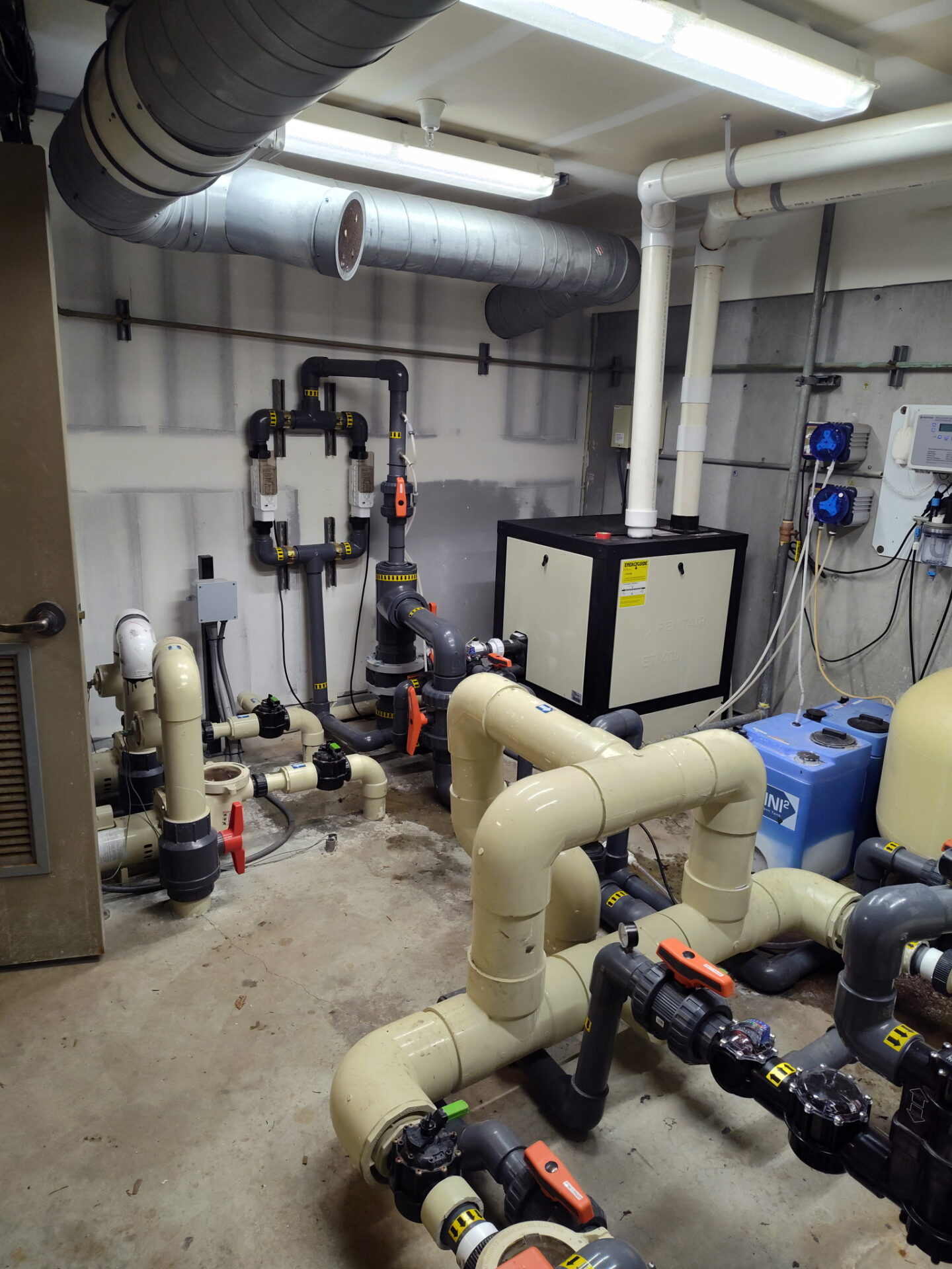 A small room filled with industrial pipes, valves, and machinery. Utilities and controls are visible, alongside connected ductwork on the ceiling.