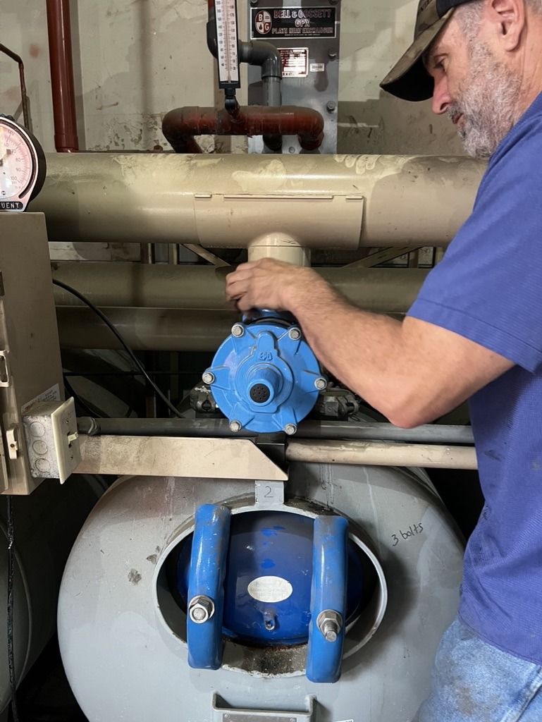 A person in a blue shirt works on a large industrial pipe system with pressure gauges and blue valves, in a mechanical setting.