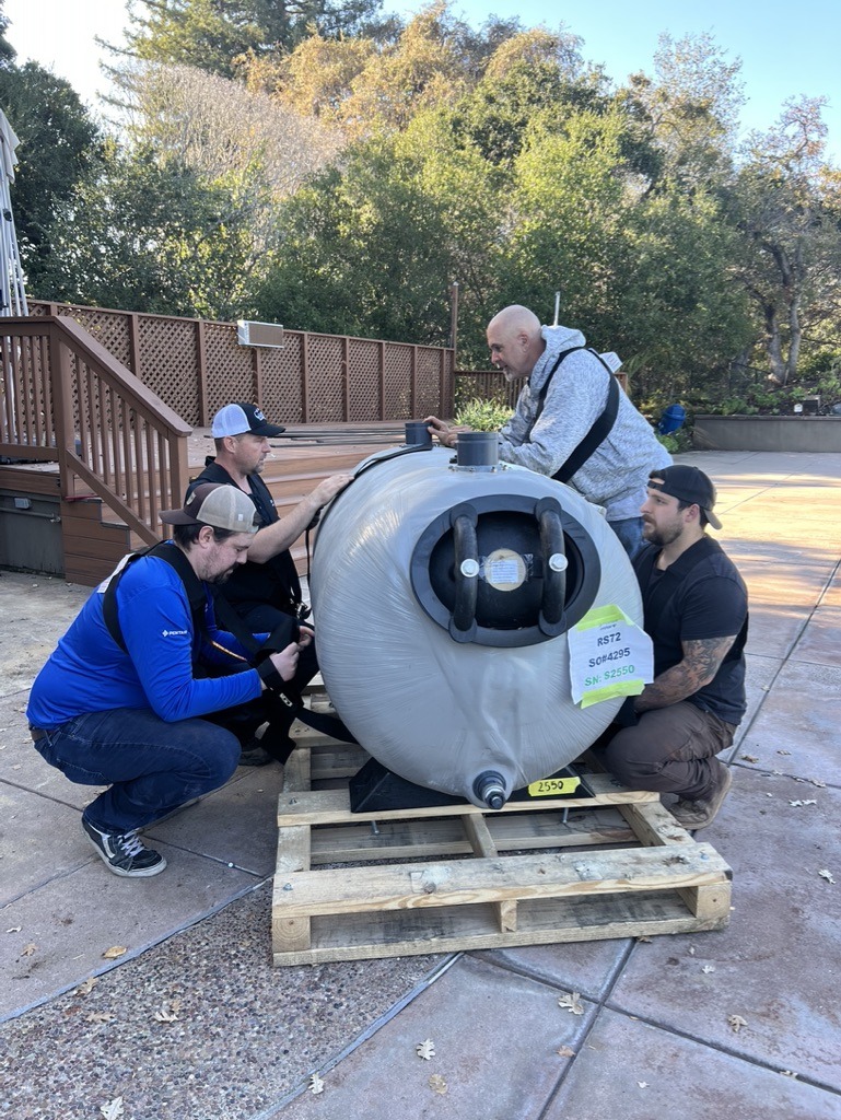 Four people examine a large, gray cylindrical object on a wooden pallet outdoors. Trees and a deck are visible in the background.