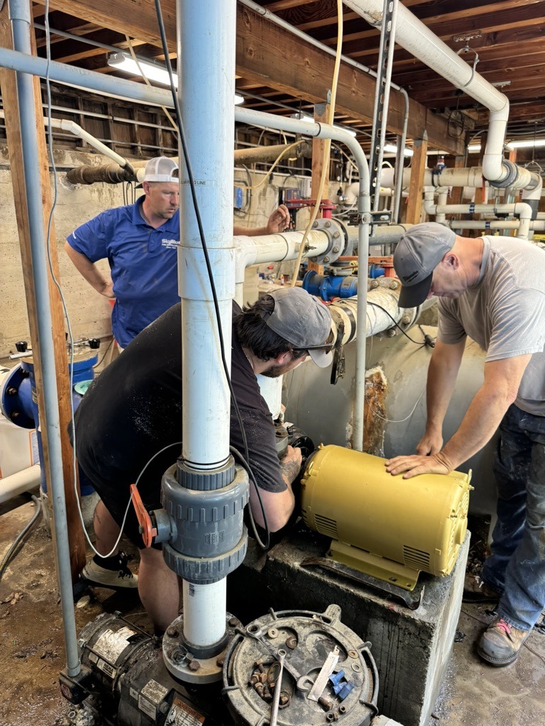 Three people work on pipes and machinery in an industrial setting. They focus on equipment repairs, surrounded by metal and plumbing components.