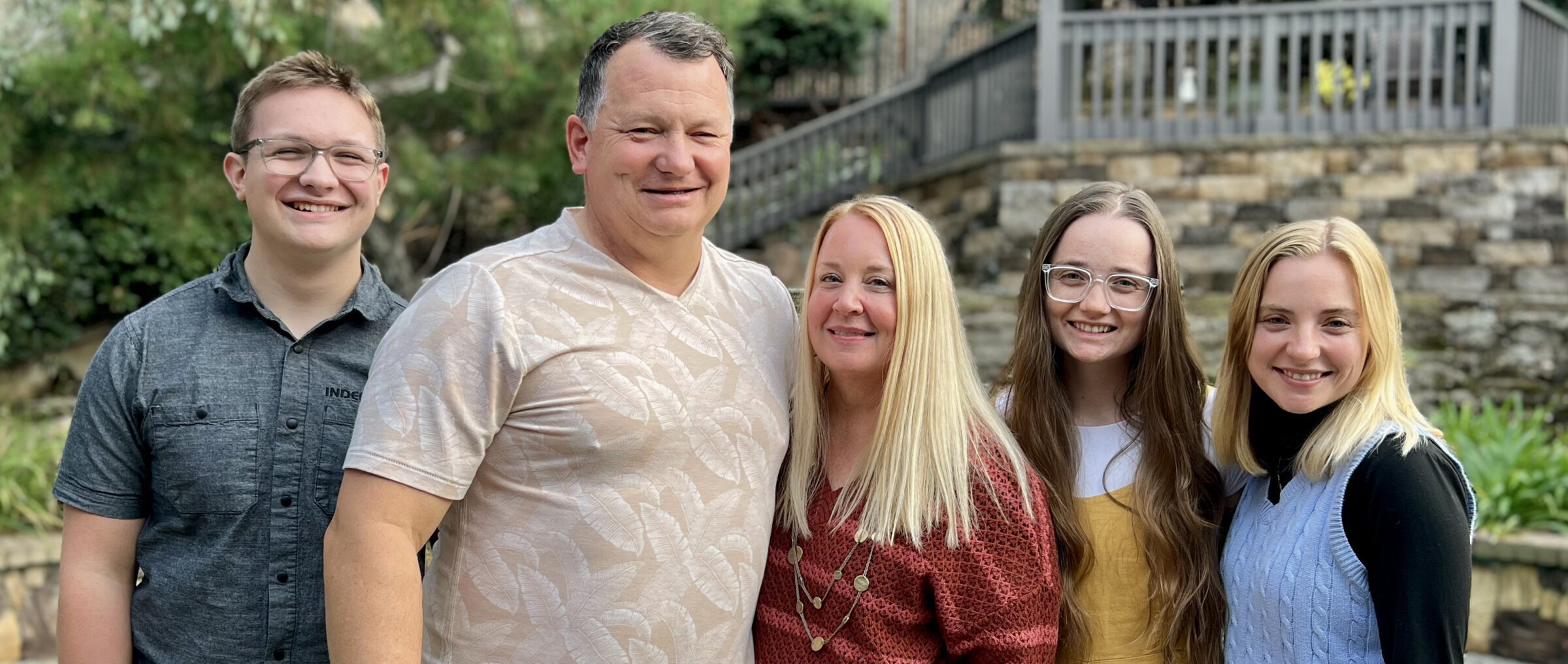 A group of five people smiling outdoors, with greenery and stone steps in the background. Casual attire suggests a relaxed, informal gathering.