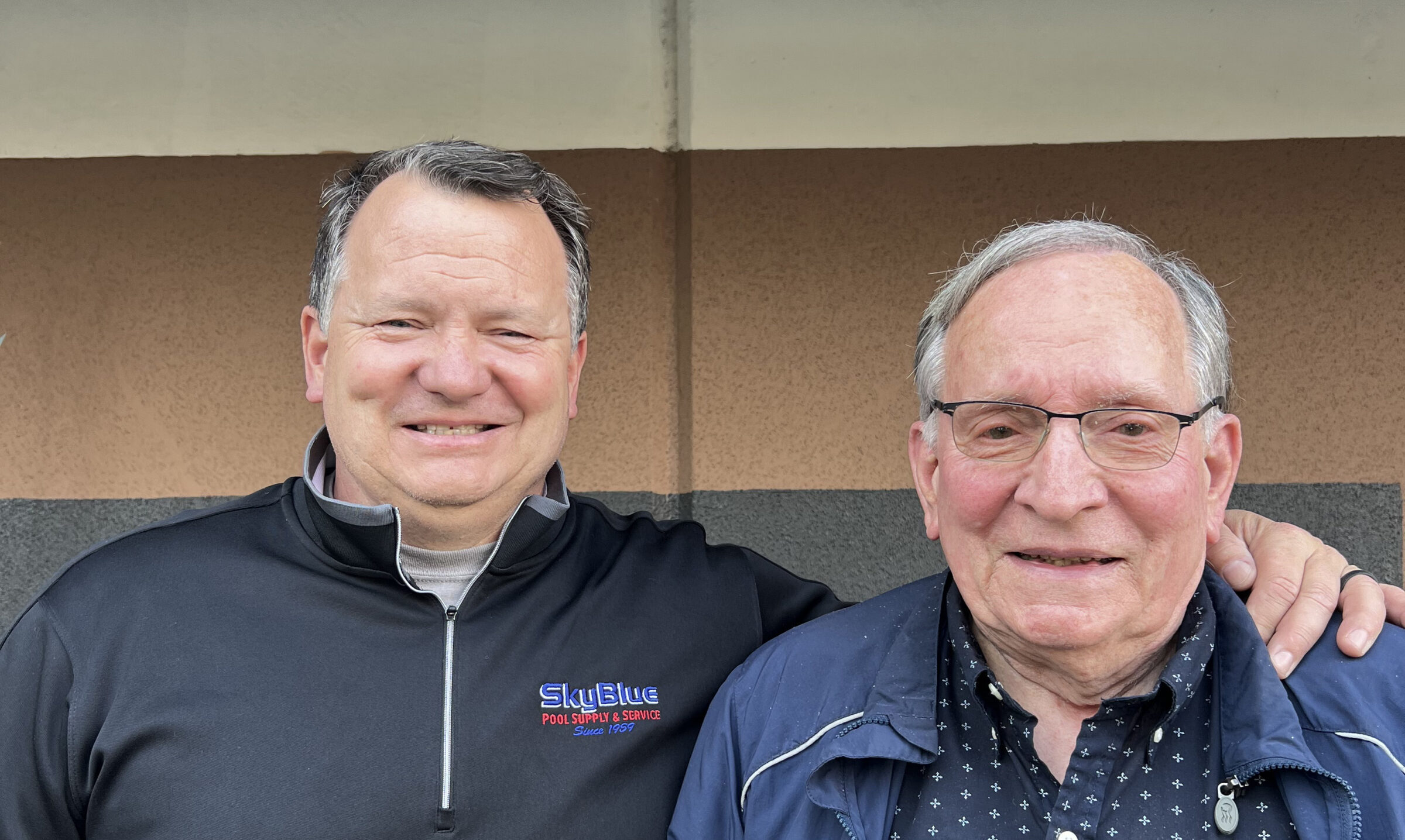 Two people smiling in front of a plain background. One person has their arm around the other. Both are wearing jackets.