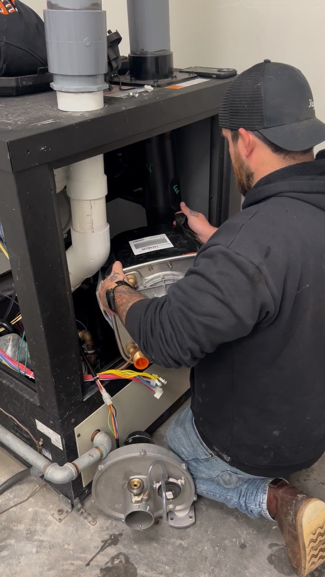 A person in a black cap repairs machinery, surrounded by pipes and wires, kneeling on a concrete floor indoors.