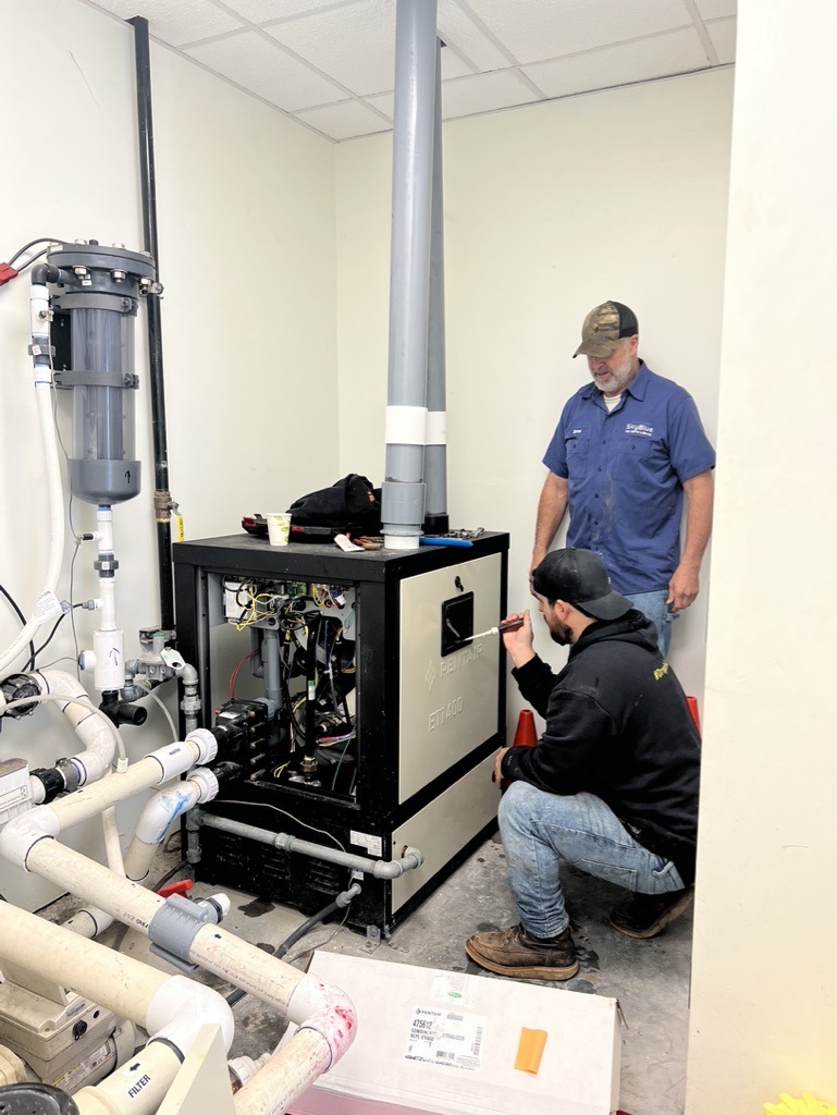 Two people inspect industrial equipment in a utility room with pipes, circuitry, and a metal cabinet. One person kneels, the other stands.