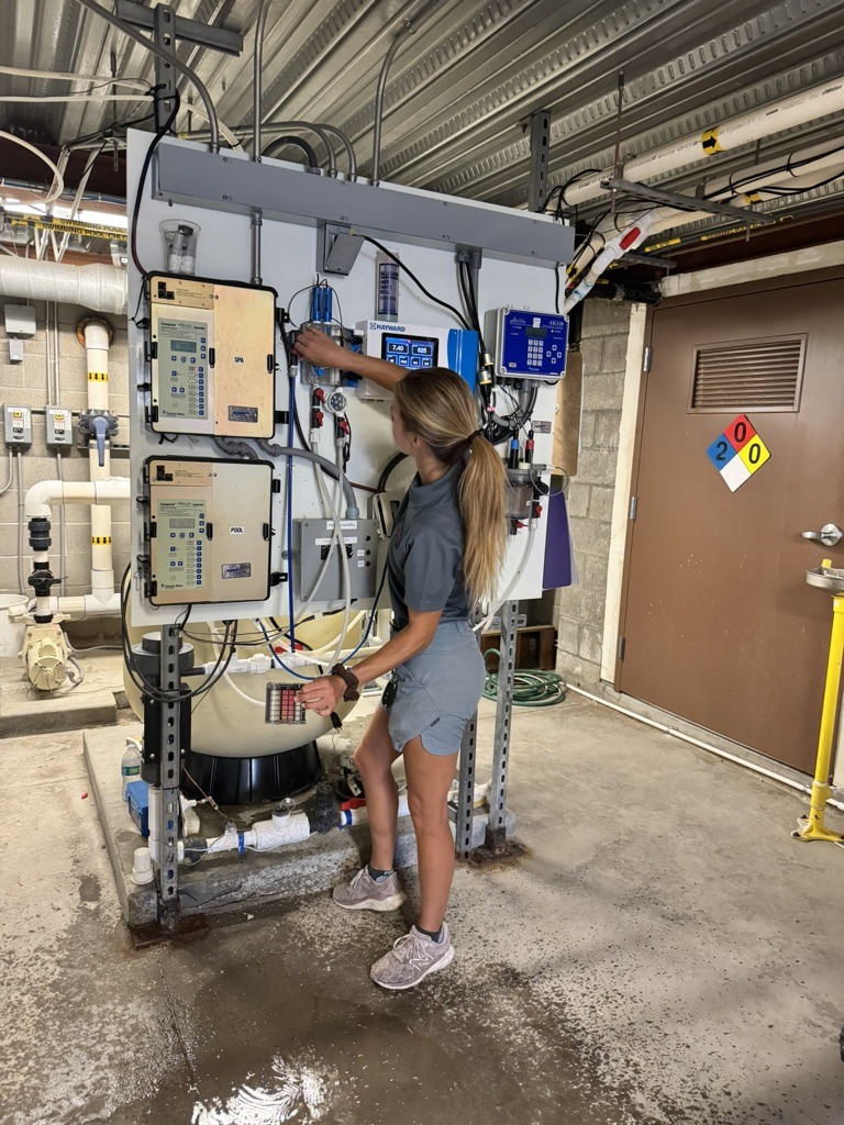 A person in a utility room operates complex equipment, adjusting controls and wiring beneath a metal ceiling, next to a labeled door.