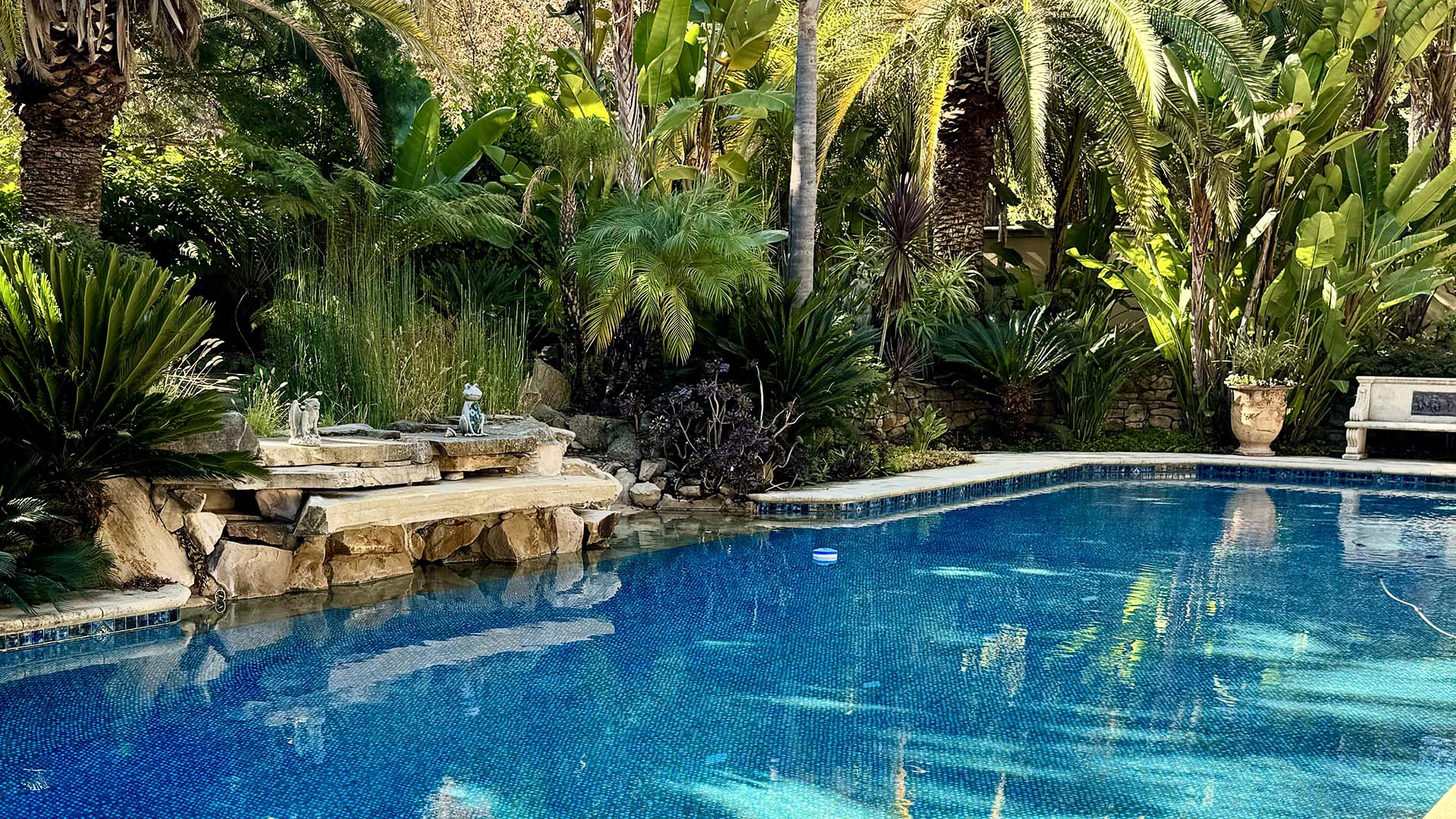 A tranquil pool surrounded by lush tropical plants and trees, featuring a stone water feature and a decorative urn.