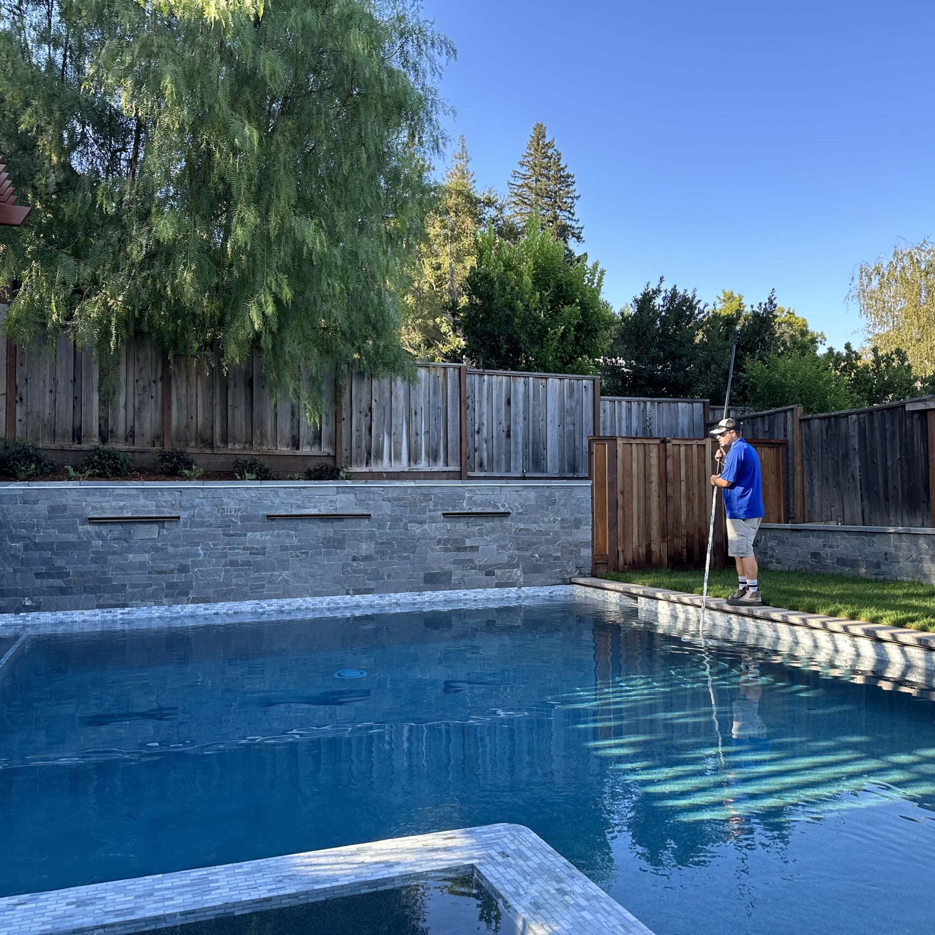 A person in blue shirt maintains a clear backyard pool surrounded by stone and wood fencing, with trees in the background under a sunny sky.