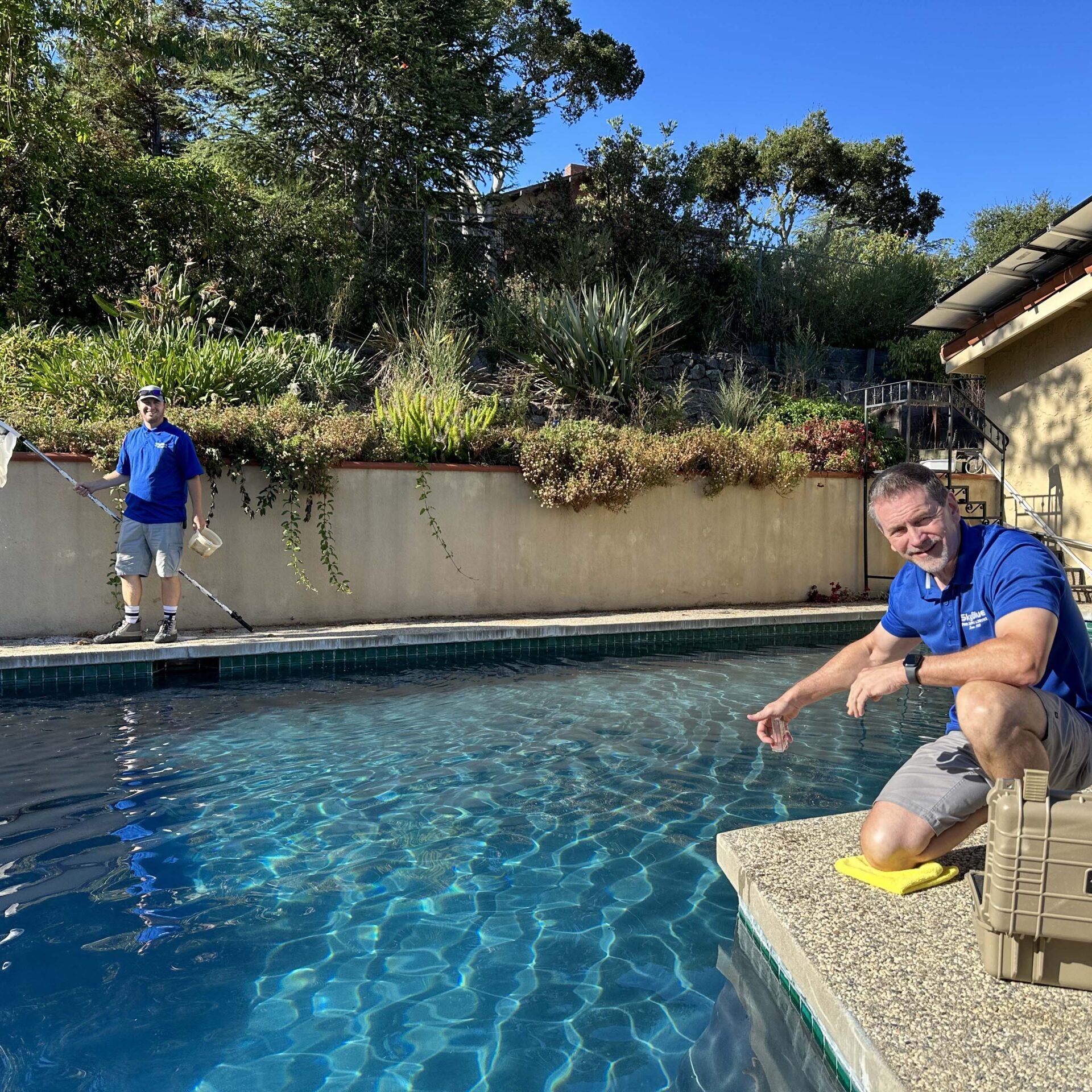 Two people by a swimming pool; one skimming water, the other pointing. Sunny day with trees and a house in the background.