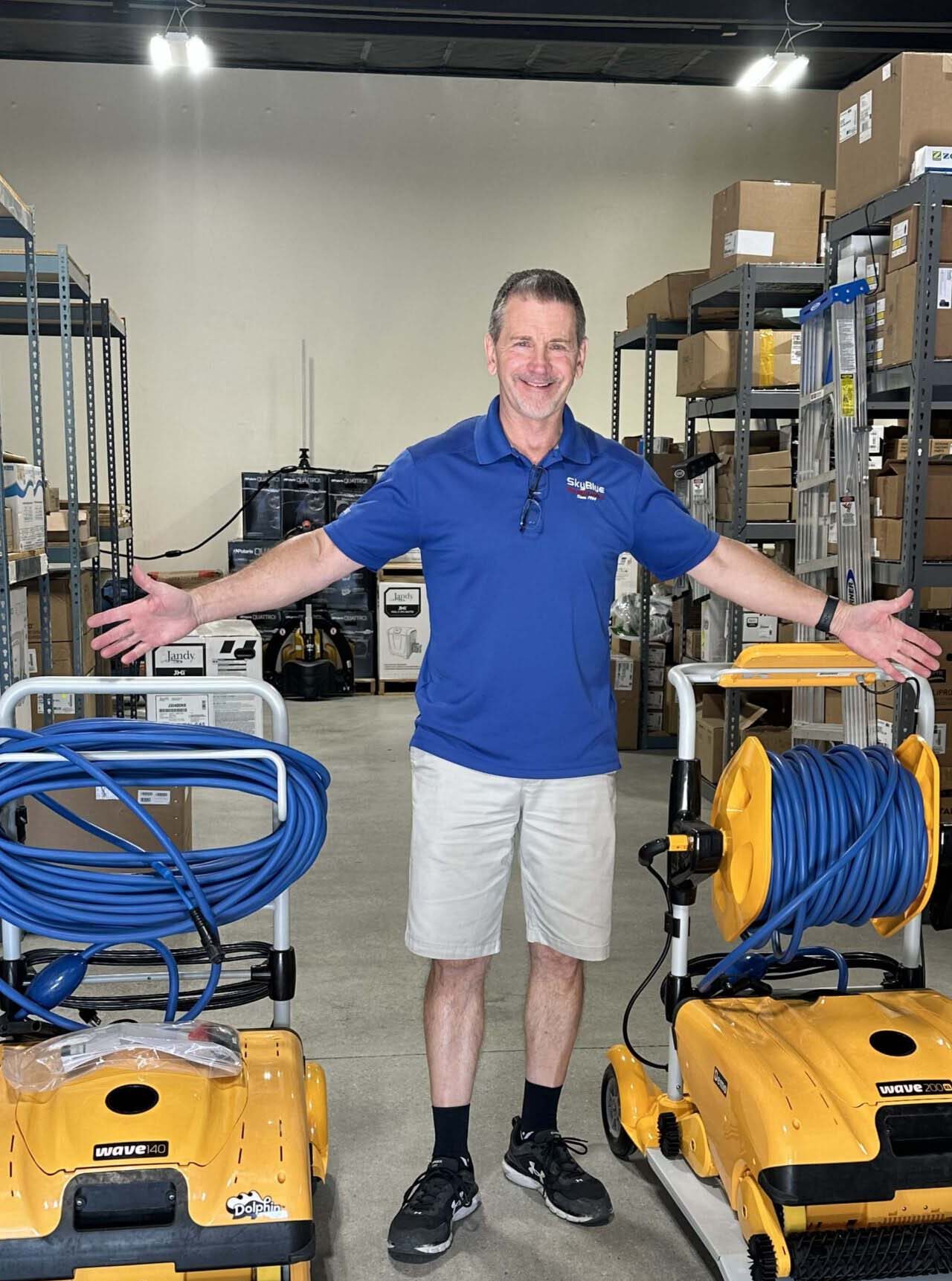 A person in a warehouse stands between two yellow industrial machines, smiling with arms open. Shelves filled with boxes and equipment surround them.