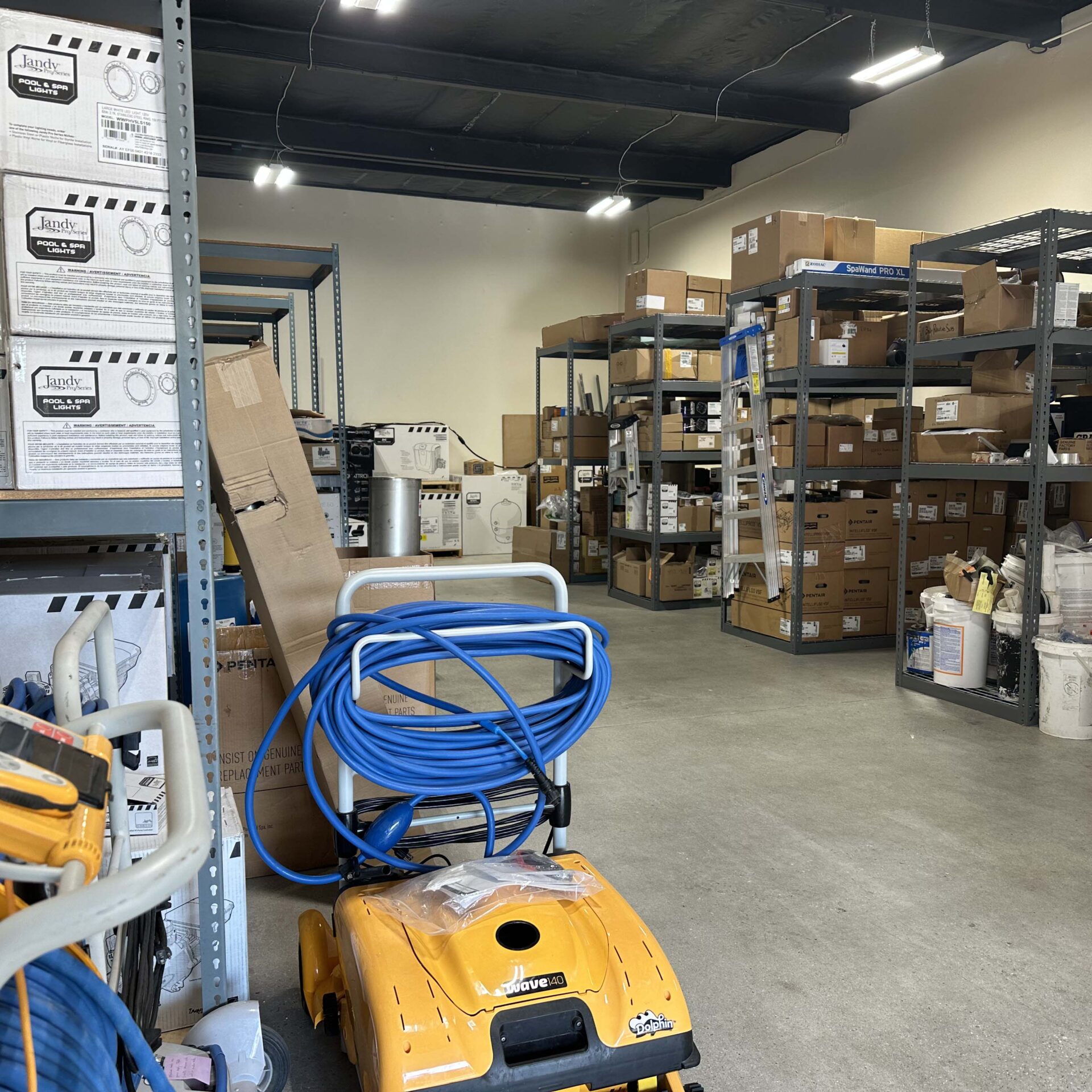 A storage room containing shelves with boxes, cleaning equipment, and various supplies is organized neatly. A ladder stands near the shelves.