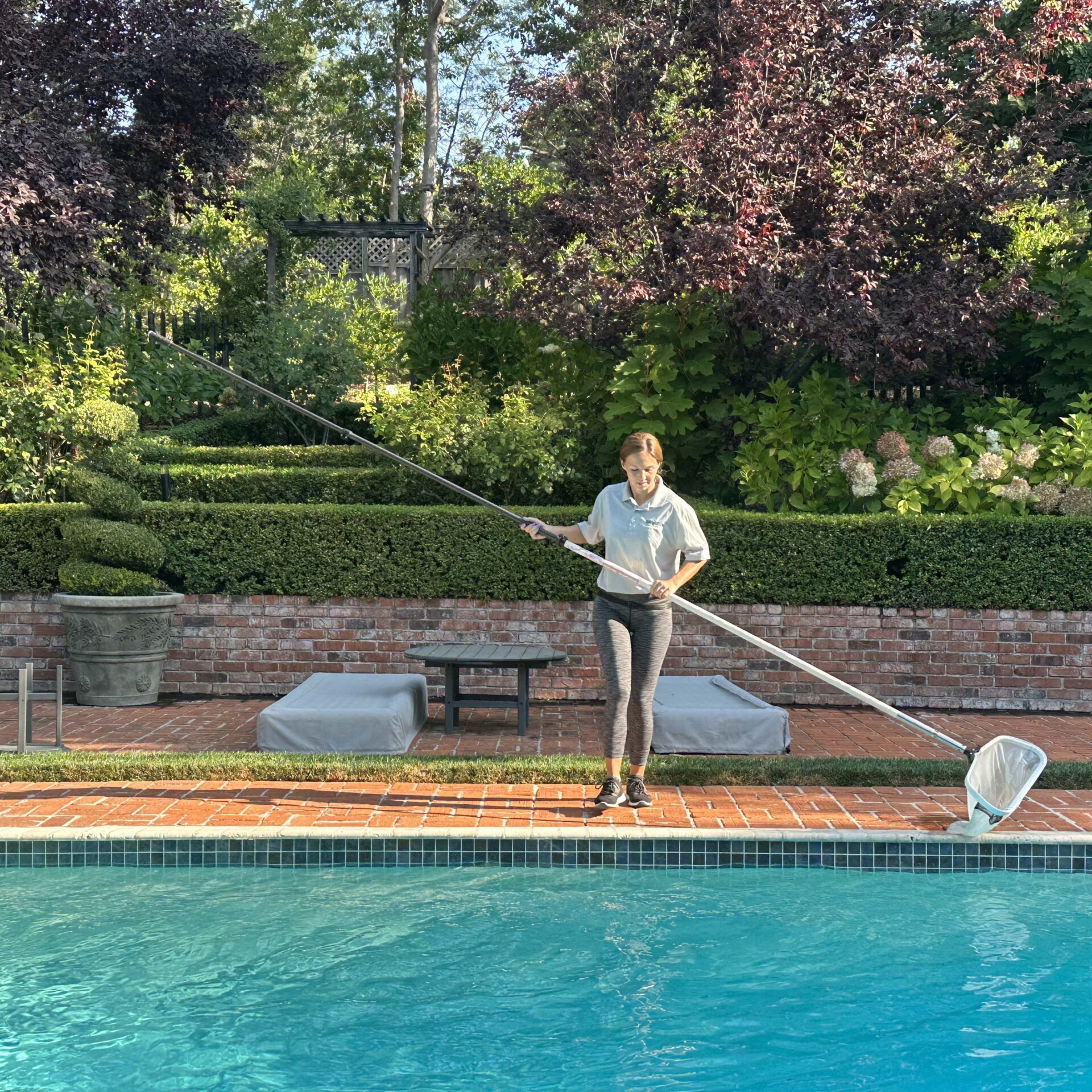 A person stands poolside, holding a long net. They're surrounded by lush greenery, neatly trimmed hedges, and brick pathways in a garden setting.