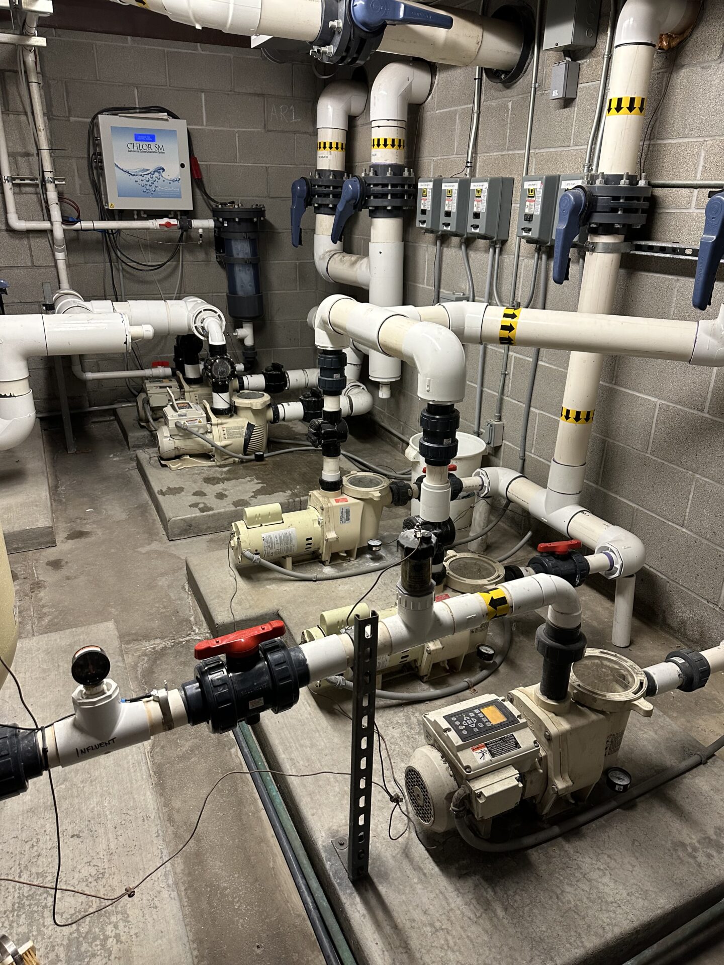 Room with multiple white pipes and pumps connected to a control panel, displaying a mechanical system setup on a concrete floor.