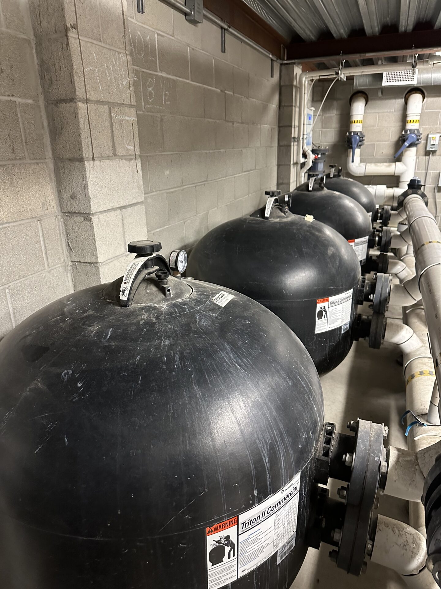 A row of large, black industrial tanks with pipes in a concrete-walled utility room, featuring gauges and warning labels.