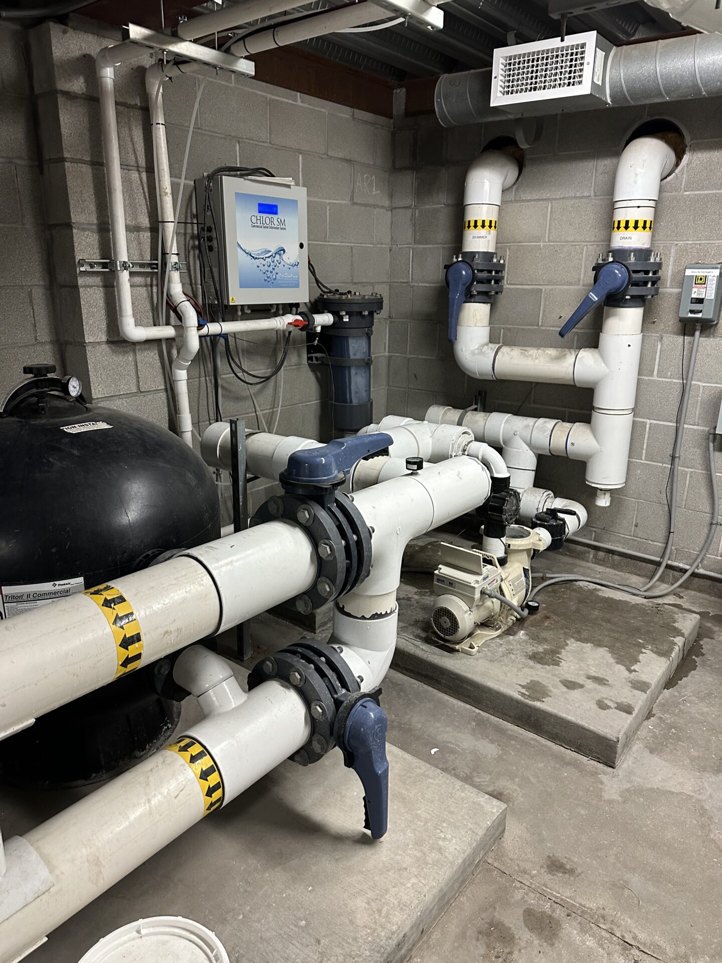 Industrial room with various pipes, valves, and machinery against a concrete wall. Equipment includes a filtration system and control panel, indicating water management.