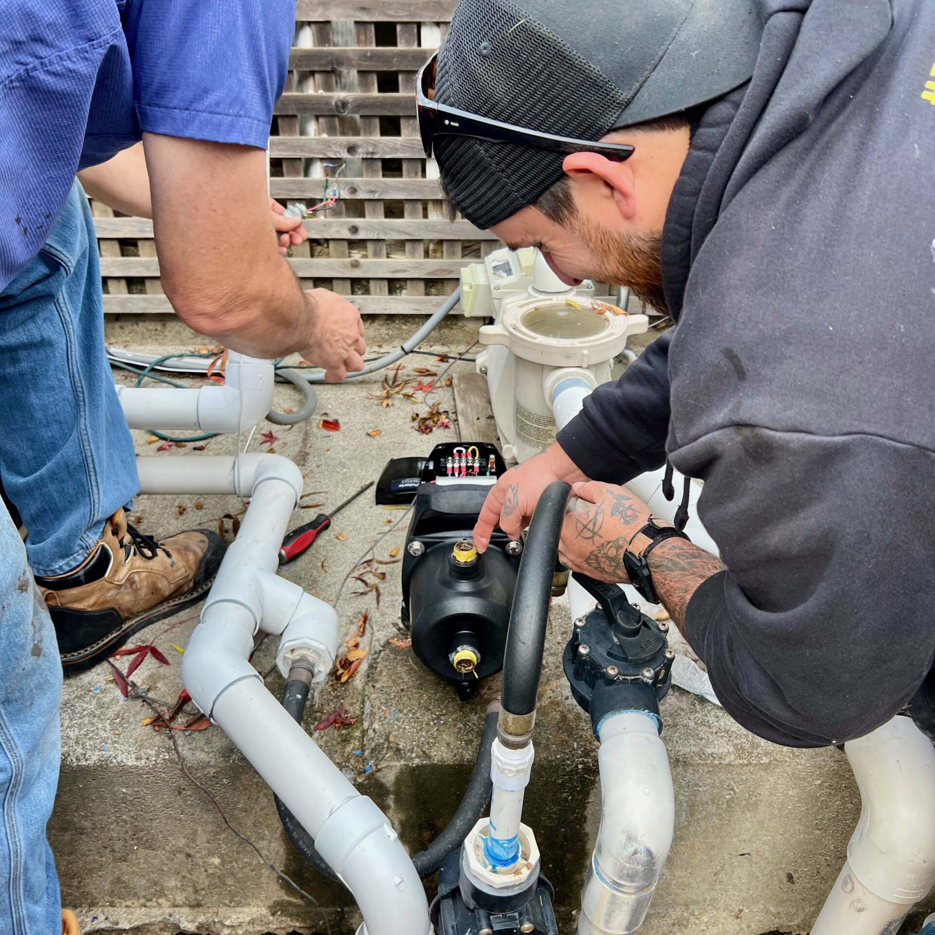 Two people are working on plumbing pipes with tools. They are focused on adjusting connections in an outdoor setting with a concrete surface.