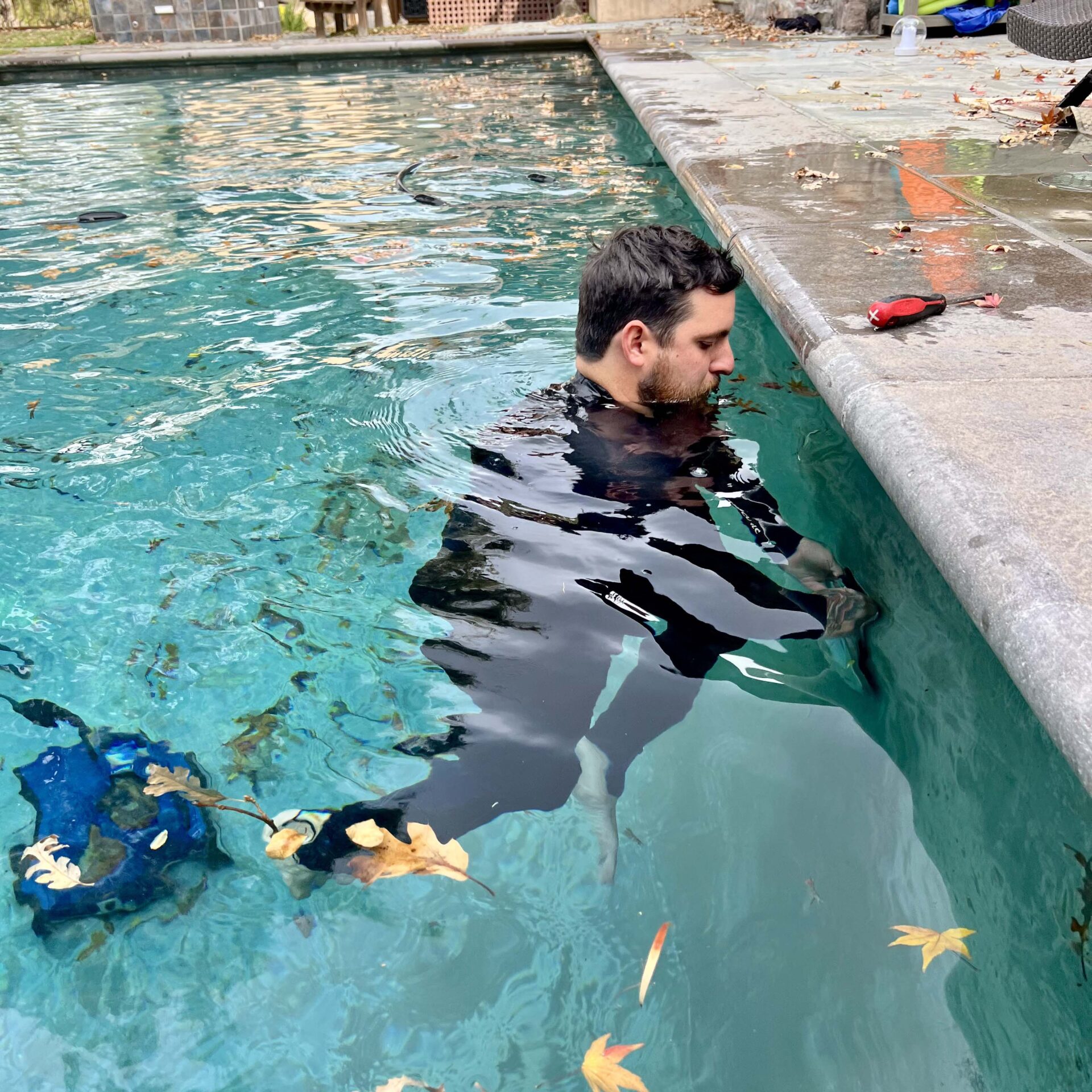 A person in a wetsuit stands in a clear swimming pool, holding a tool. Leaves and equipment are visible on the surface and deck.