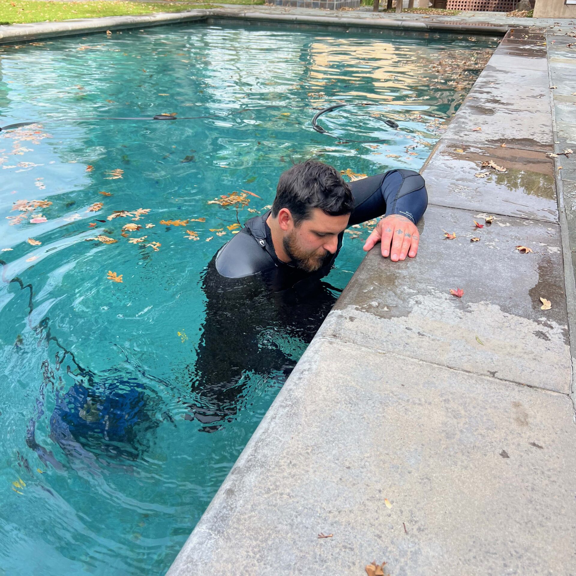 A person in a wetsuit is in a leaf-filled pool, holding onto the edge, with green grass visible in the background.