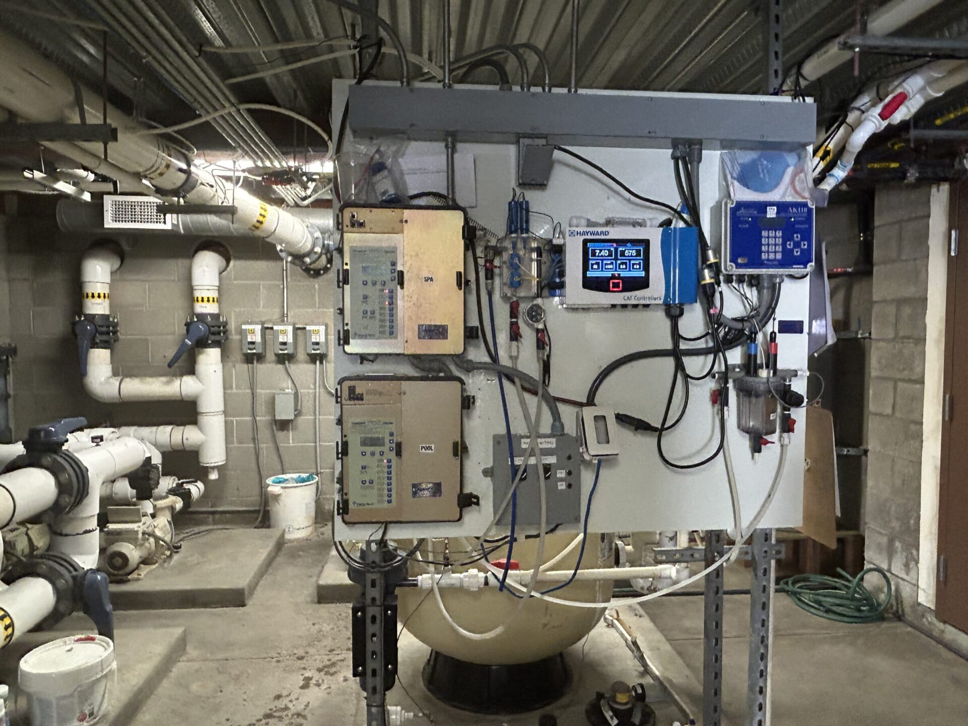 Industrial control room featuring various machines, panels, and pipes. The setting includes complex wiring and plumbing, suggesting a technical or utility-related purpose.