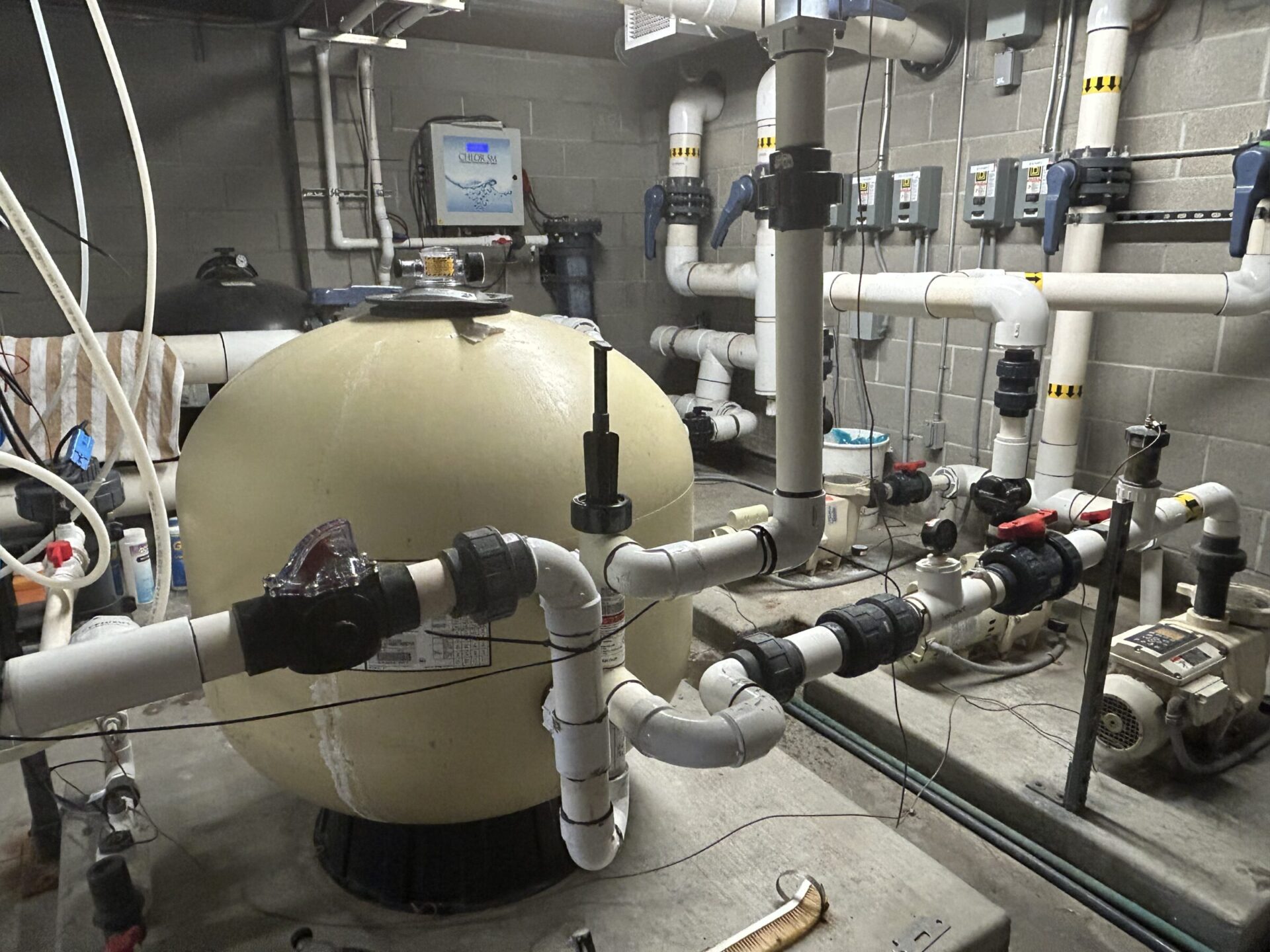 A complex pipe system in a utility room features a large filtration tank, various valves, gauges, and control equipment on the walls.