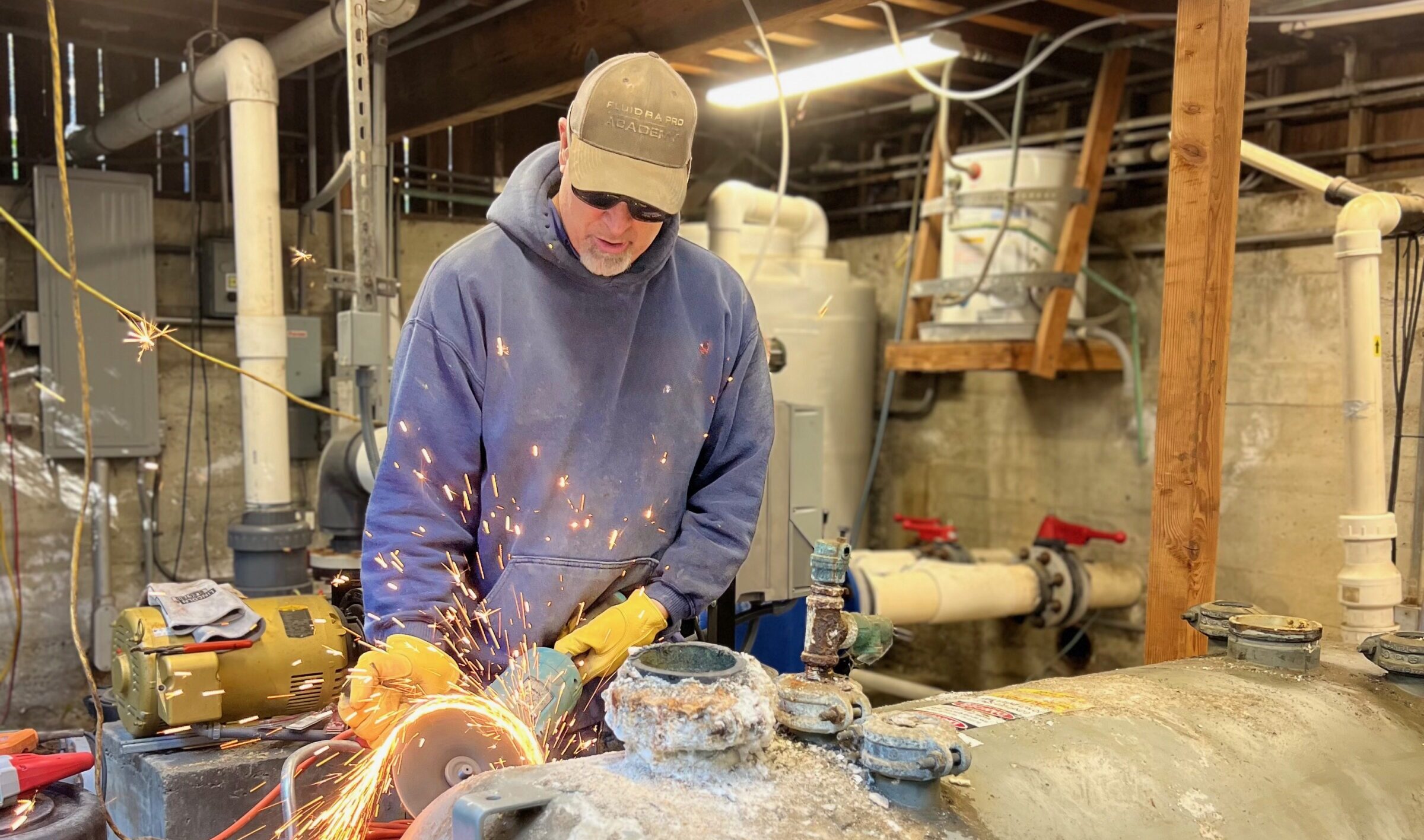 A person wearing a cap and safety gloves uses a grinder on metal, creating sparks, in an industrial workshop setting.