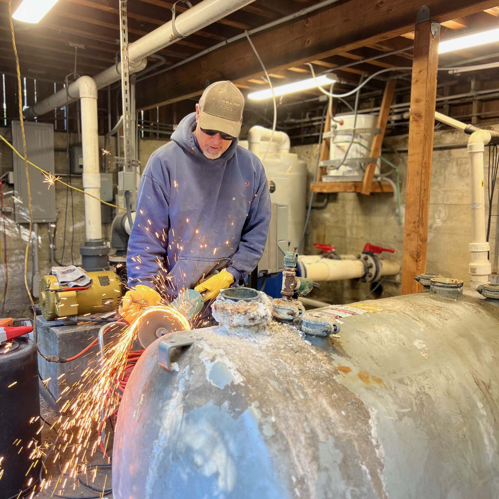 A person wearing safety gear uses an angle grinder, creating sparks in an industrial setting with pipes and machinery in the background.