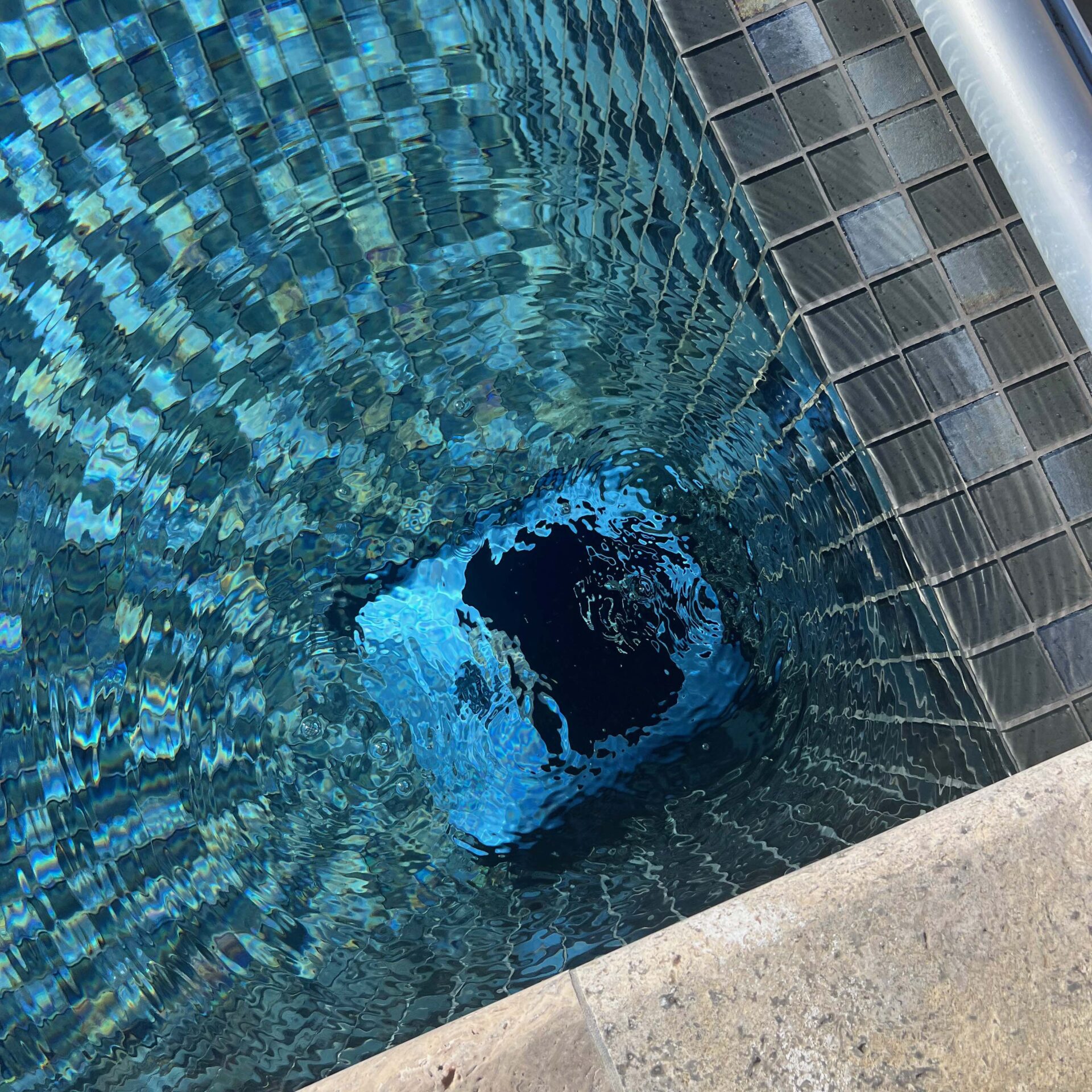 Clear swimming pool with a shaded square opening at the bottom, surrounded by light-colored tiles, reflecting sunlight on rippling water.