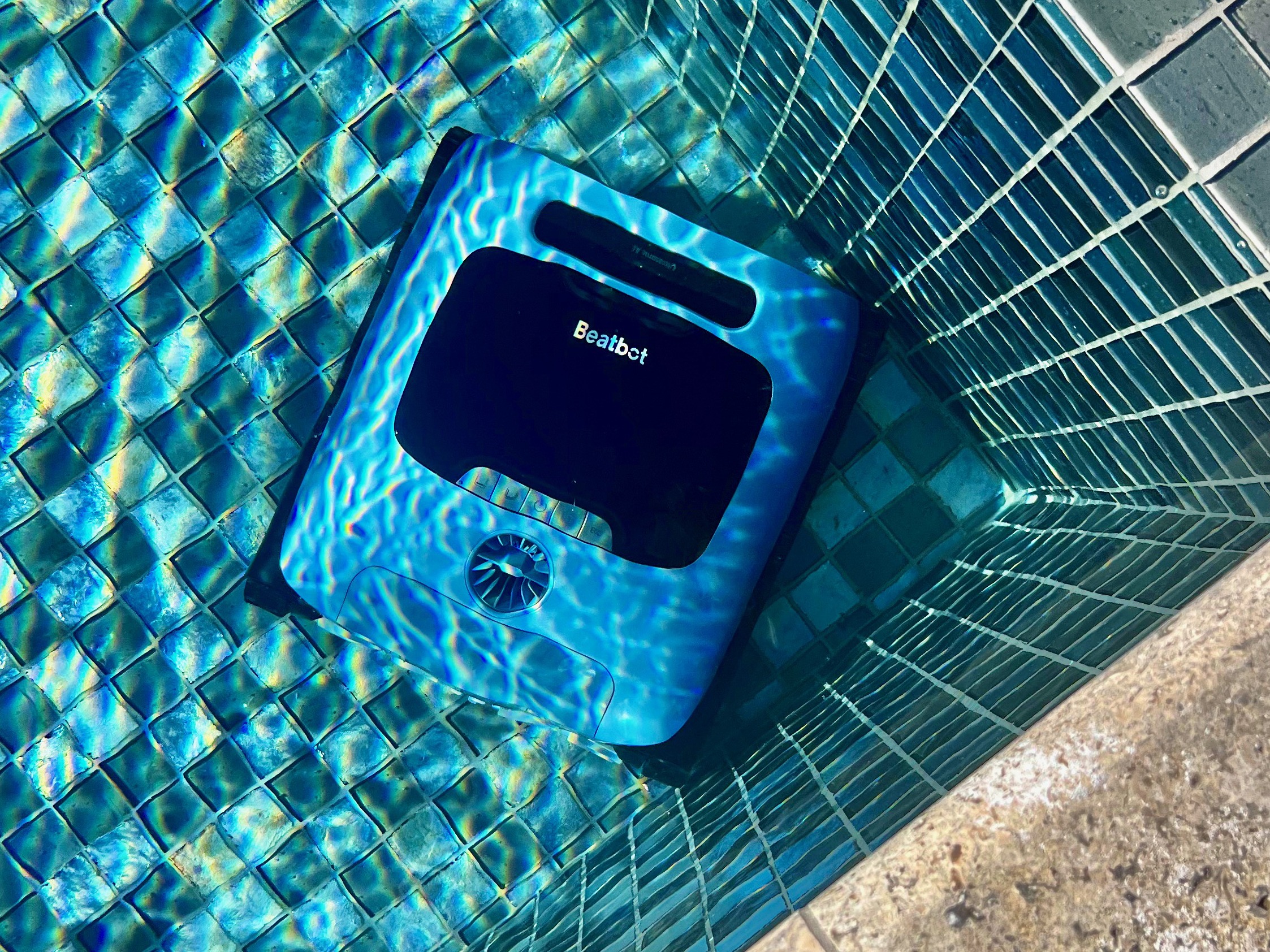 A pool cleaner named Seabot rests at the bottom of a tiled pool, with clear blue water casting reflections on its surface.