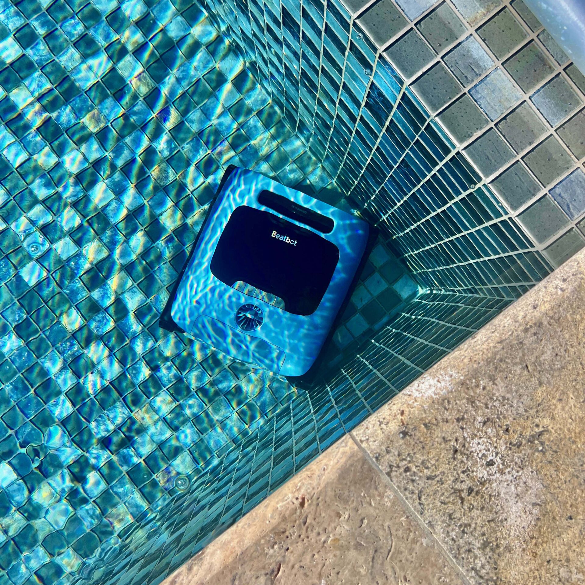 Pool cleaning robot submerged in a brightly lit swimming pool, with mosaic tile design visible. Sunlight creates shimmering water reflections.