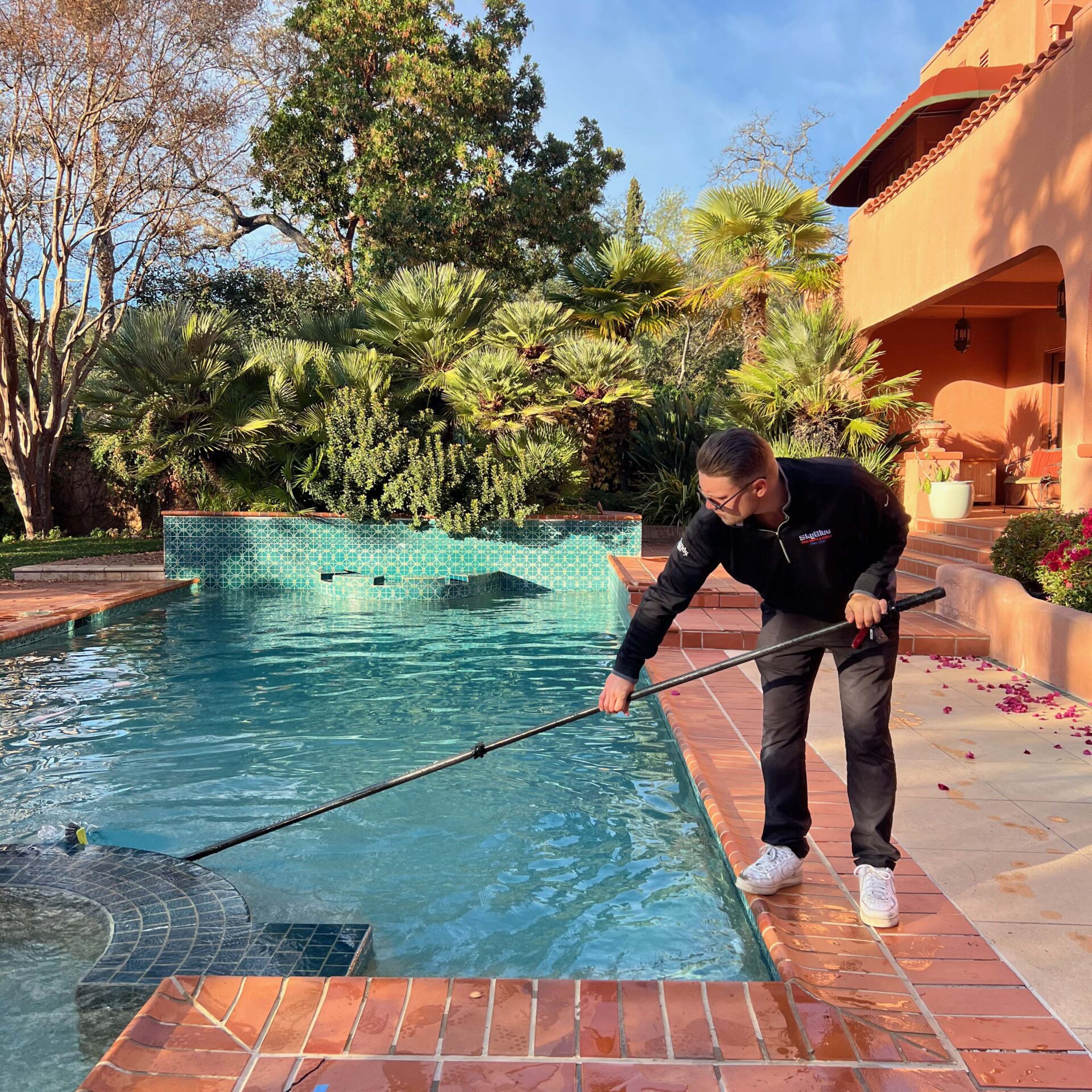 A person is cleaning a swimming pool with a long pole in a tropical backyard setting beside an orange building under a clear sky.