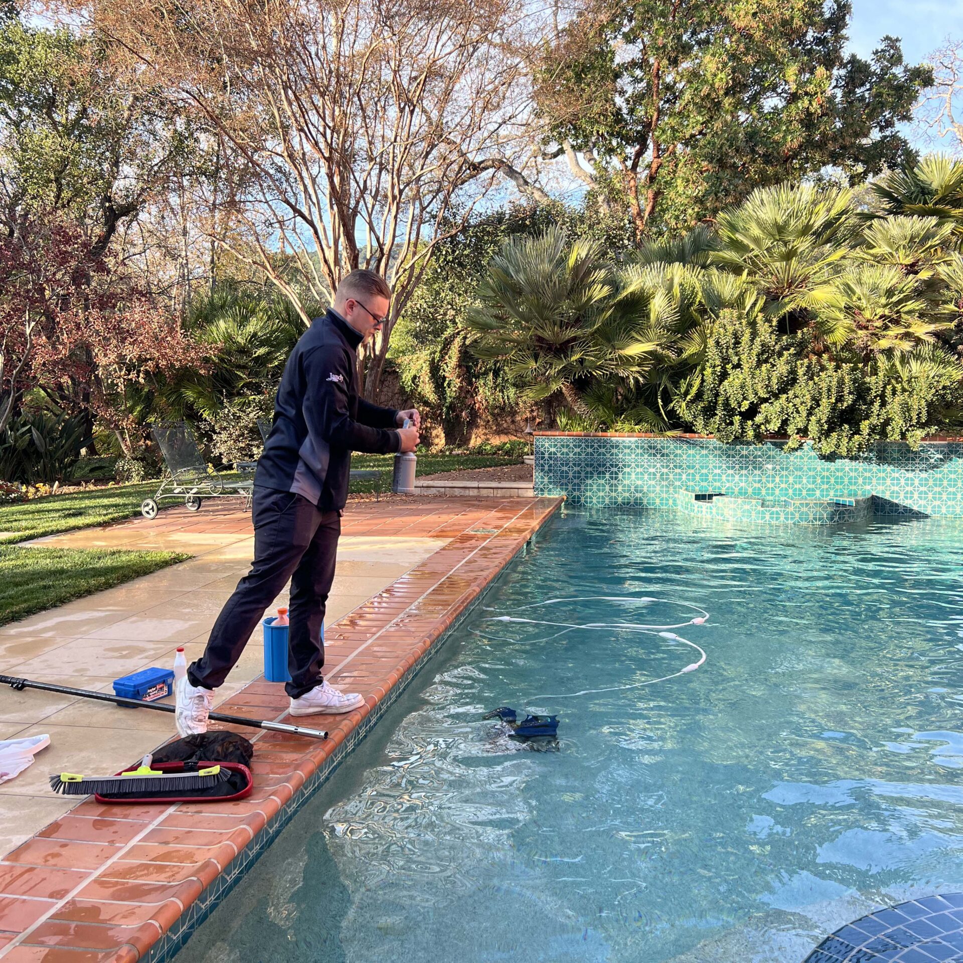 A person stands near a swimming pool testing water, surrounded by lush greenery and a serene outdoor setting.