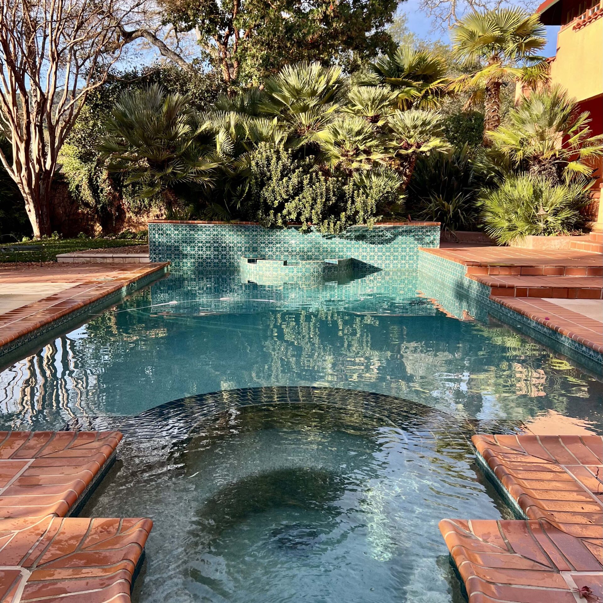 A serene swimming pool with turquoise tiles, surrounded by lush greenery and terracotta accents, reflecting a tranquil outdoor setting under clear skies.