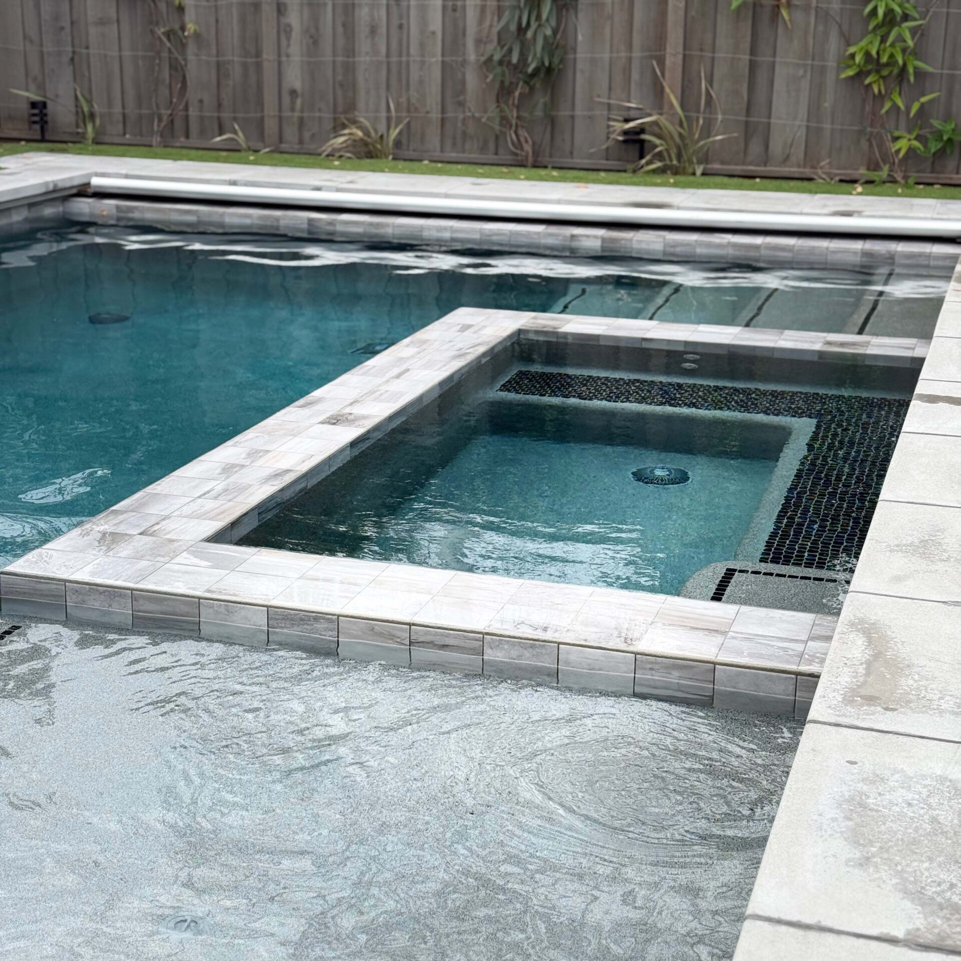 A modern, rectangular pool with a small hot tub. Surrounding tiles are light gray. Wooden fence and sparse greenery in the background.