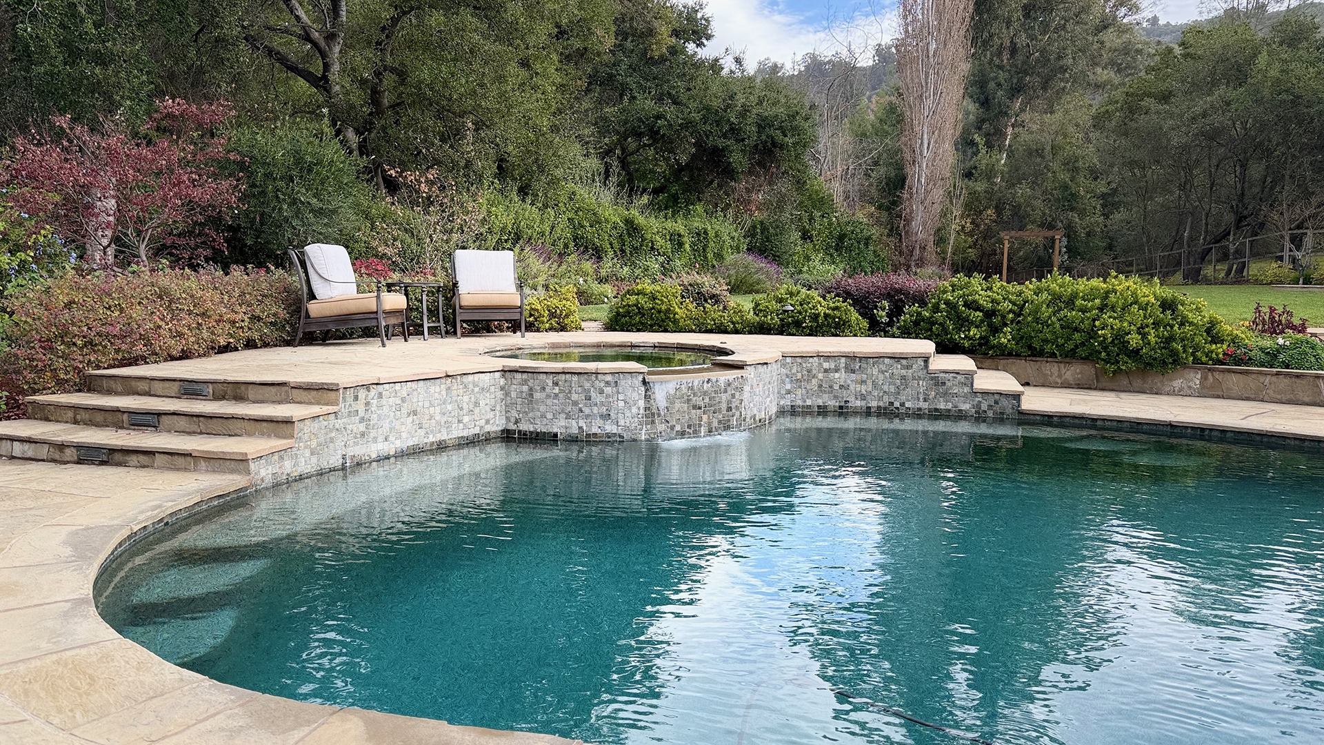 A serene backyard features a swimming pool, hot tub, lush greenery, and two patio chairs, creating a relaxing outdoor retreat.