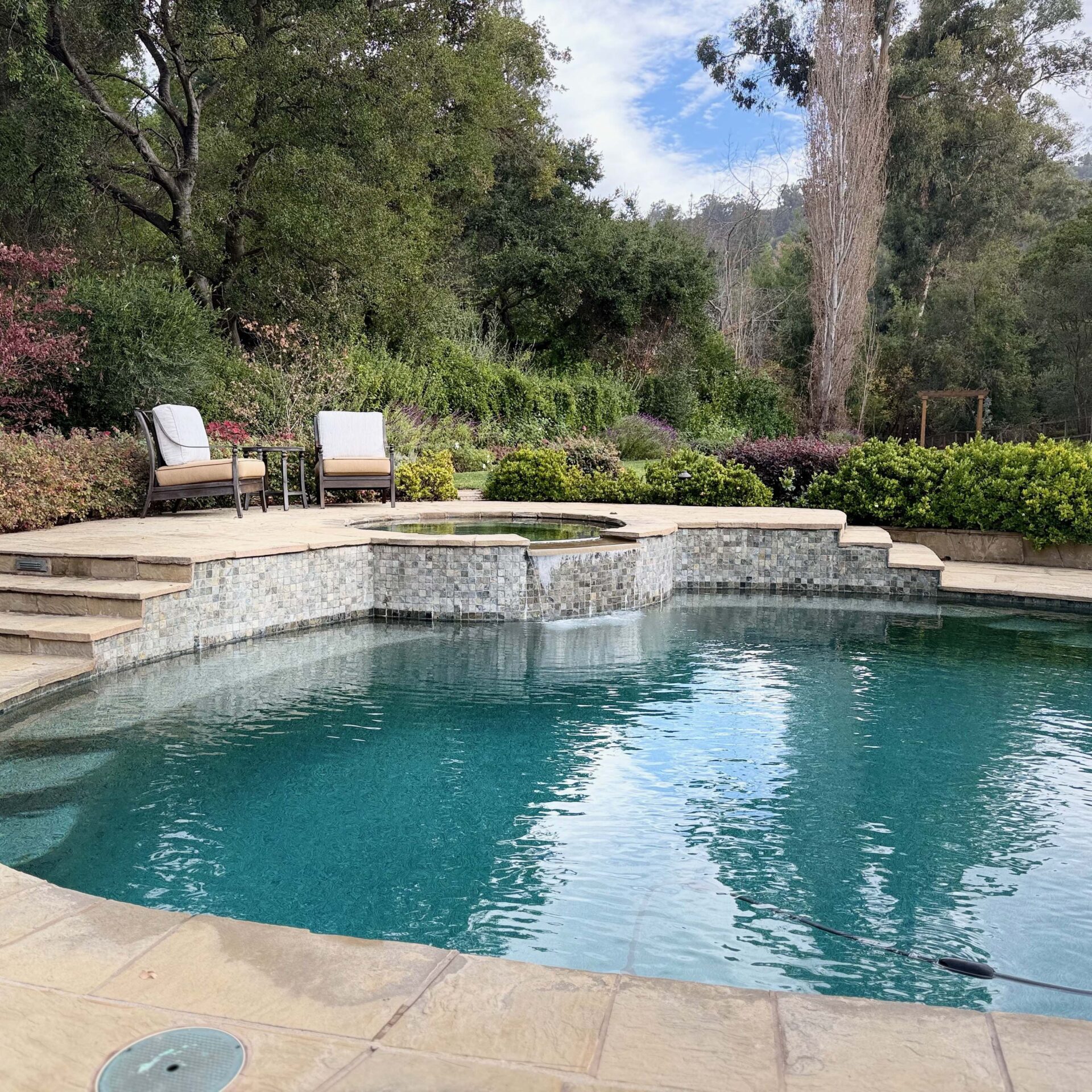 A serene backyard pool with seating, surrounded by lush trees and greenery, providing a peaceful and inviting outdoor space for relaxation.