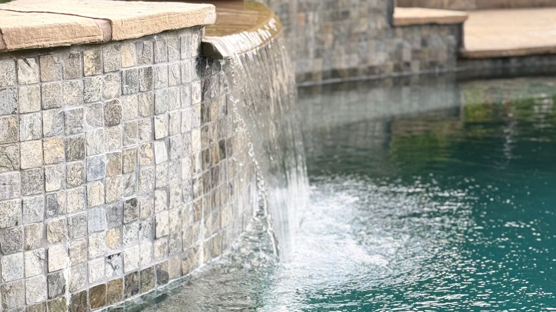 A serene pool with a stone-tiled waterfall edge, surrounded by lush greenery, offers a tranquil and inviting atmosphere.