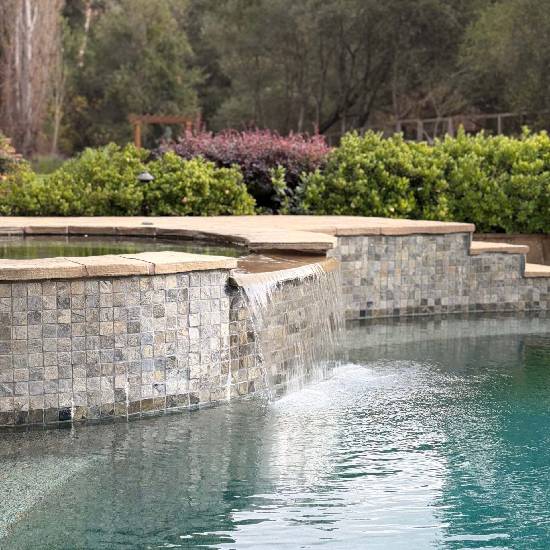 A serene pool with a waterfall feature, surrounded by lush greenery and trees, creates a peaceful outdoor setting.