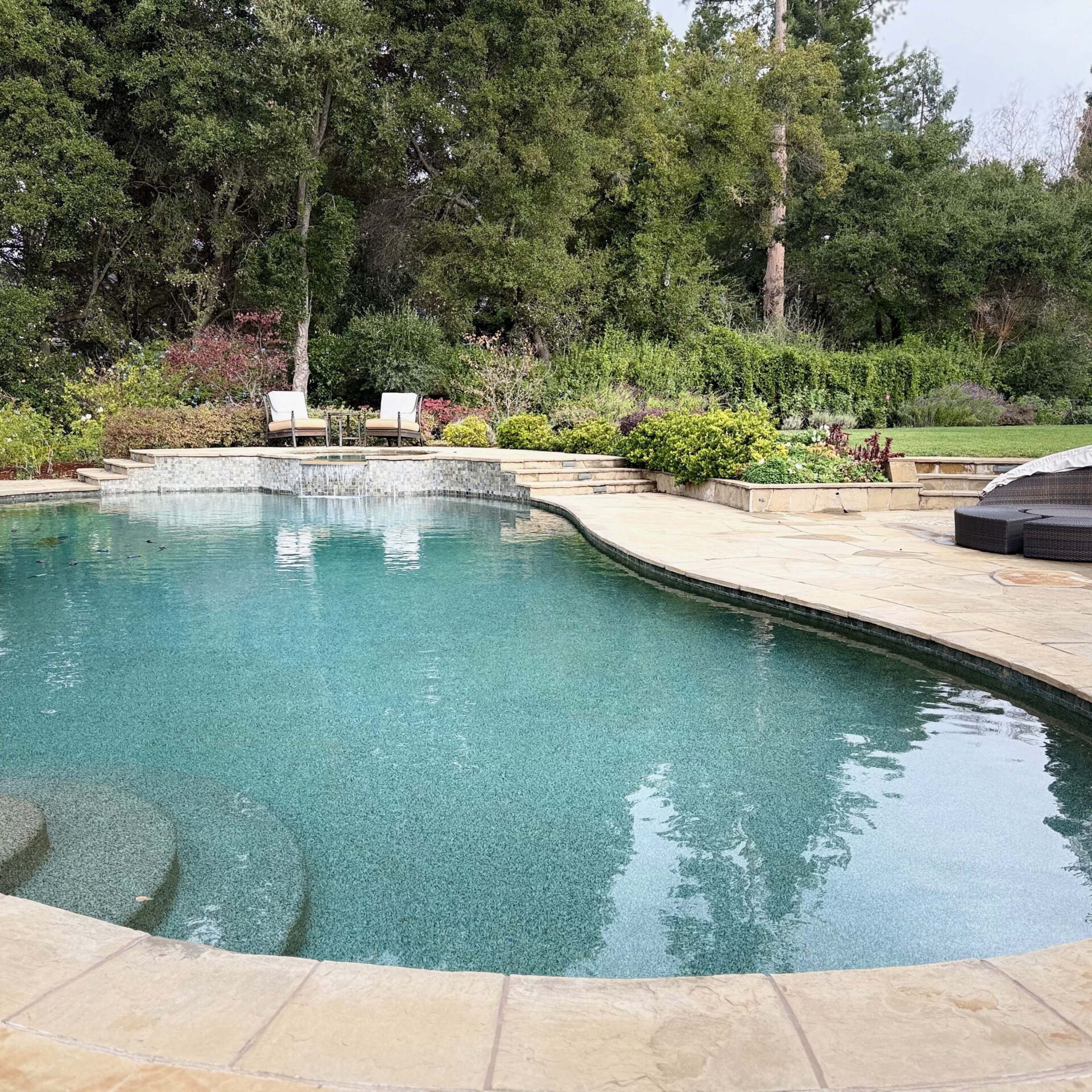 A tranquil backyard features a kidney-shaped pool, surrounded by lush greenery and lounge chairs on a stone patio under a cloudy sky.