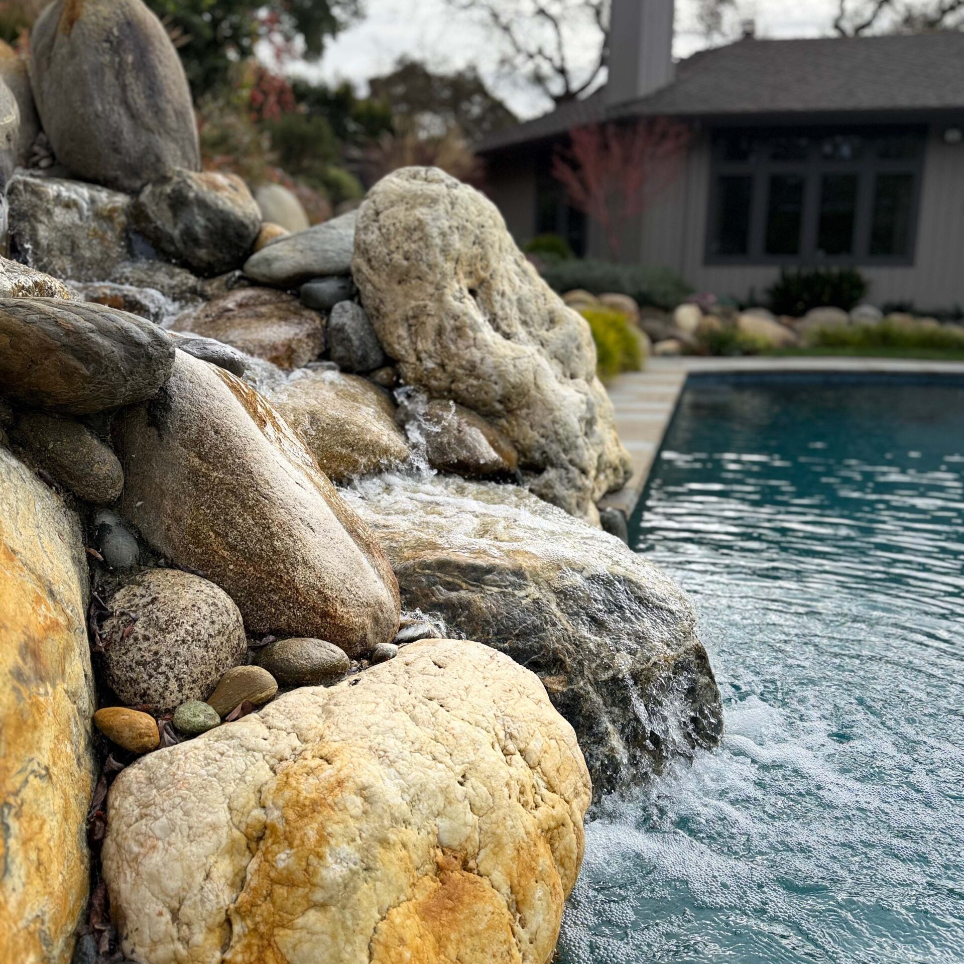 Rock waterfall beside a clear swimming pool with a house in the background. Natural landscaping creates a serene, tranquil outdoor scene.