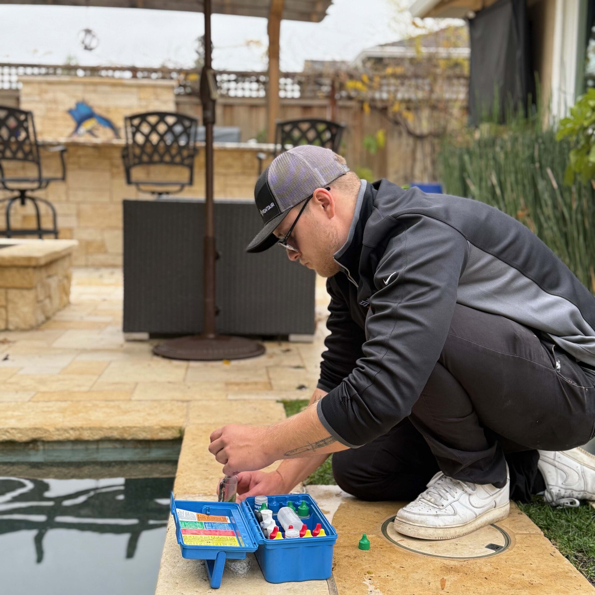 A person crouches by a swimming pool with a kit, possibly testing water quality, in a backyard patio setting.