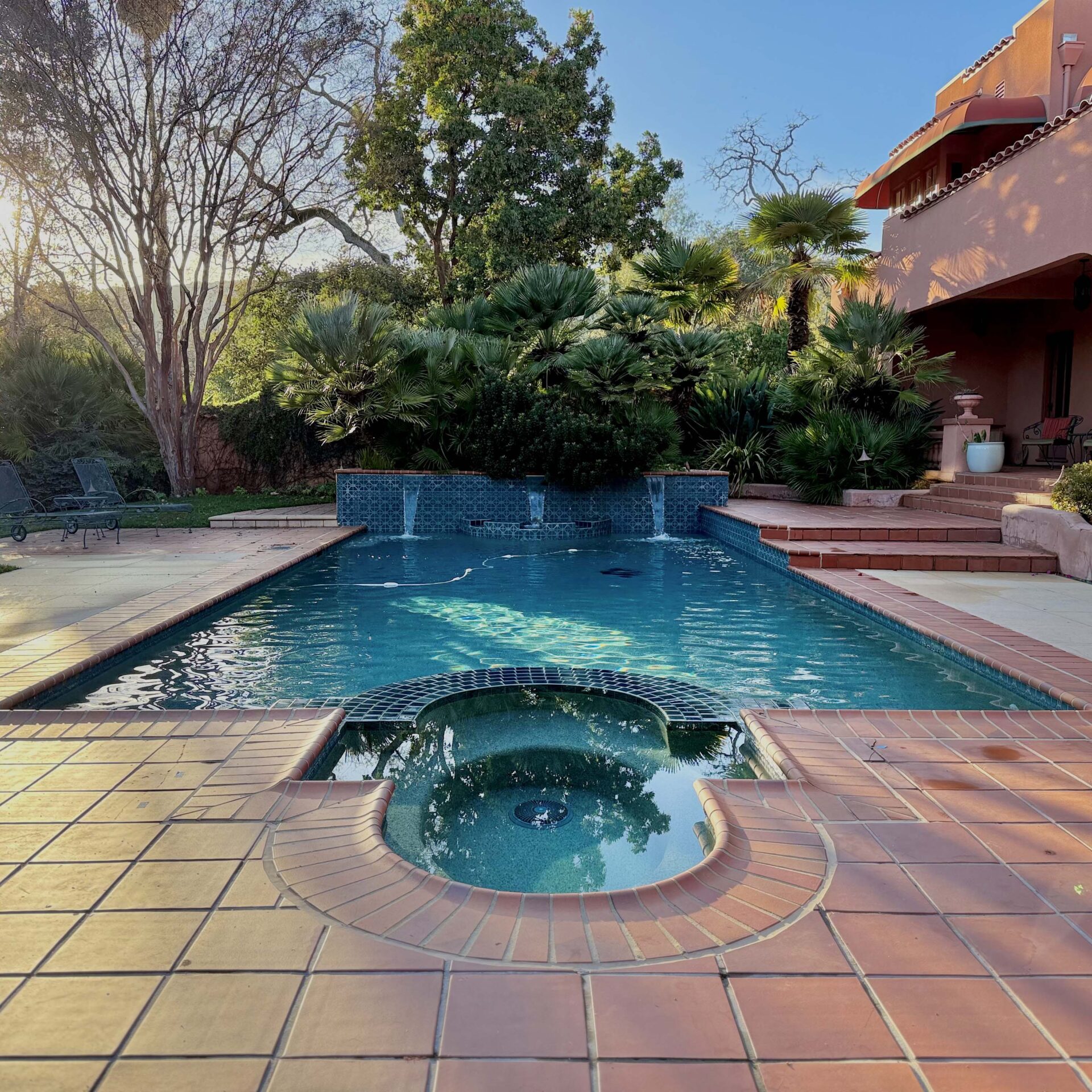 A serene swimming pool with a tile patio, surrounded by lush greenery and adjacent to a warm-toned house in a sunny landscape.