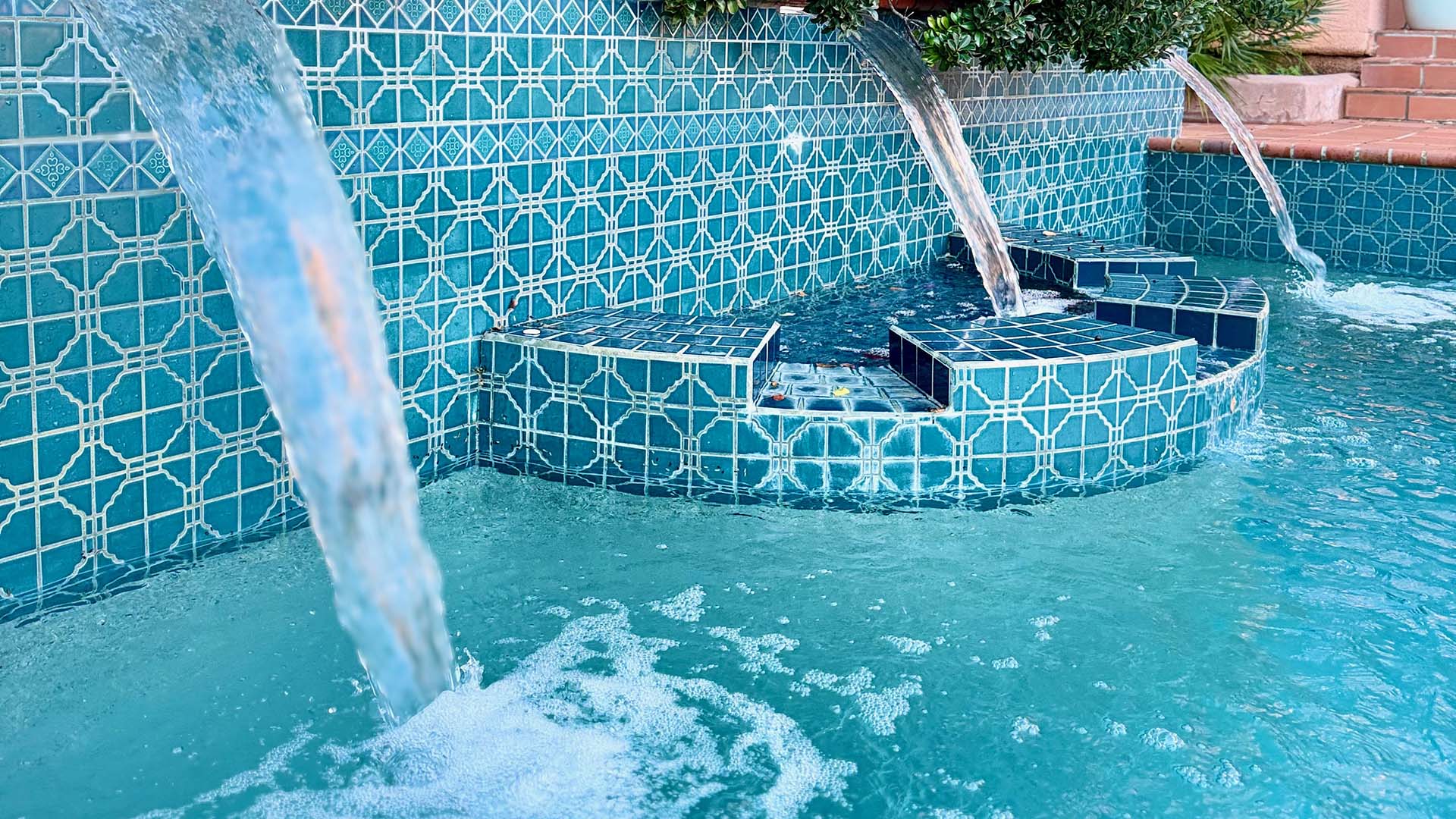 A close-up of a decorative, tiled water fountain with multiple streams, featuring geometric patterns and light blue water. No people or landmarks visible.