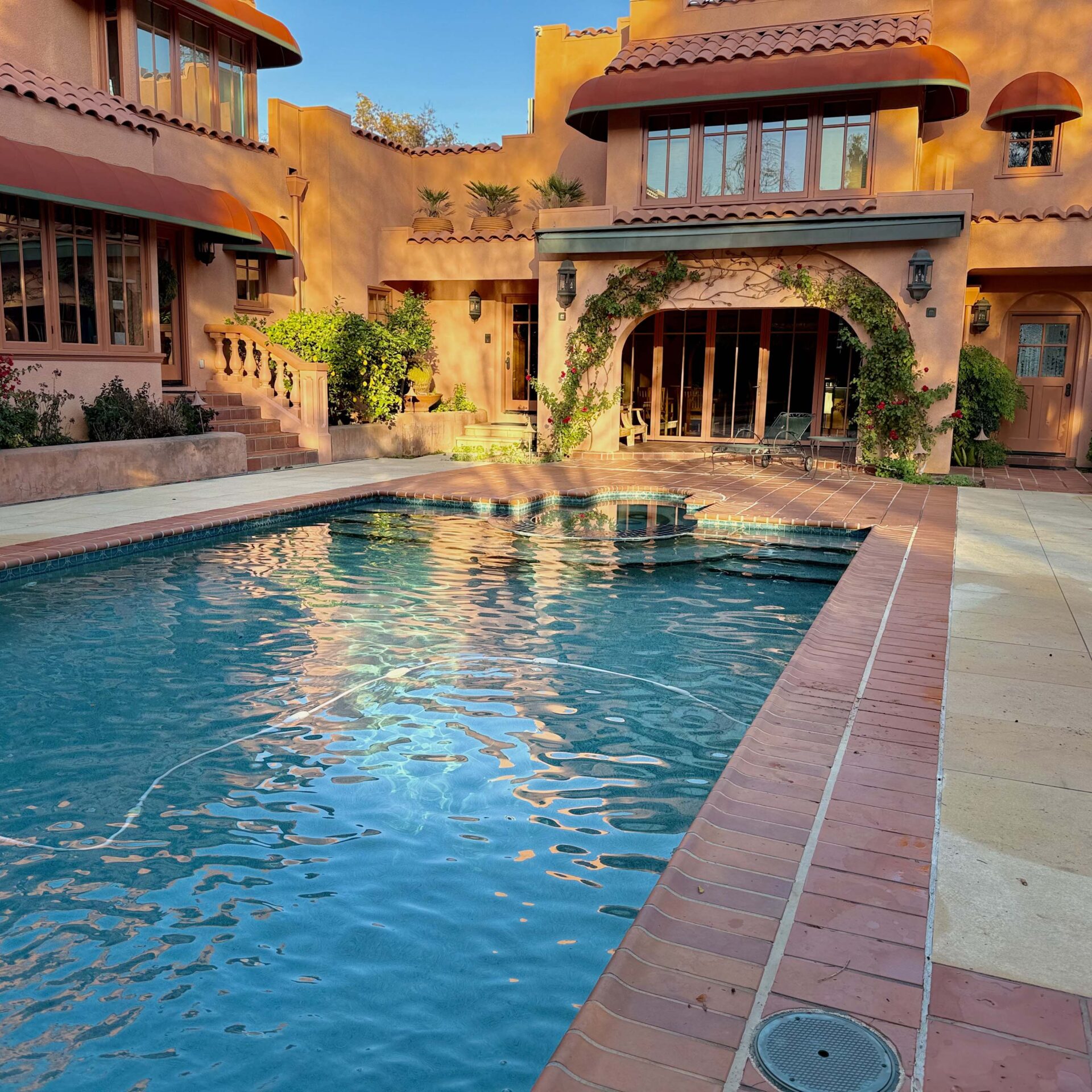 An elegant Mediterranean-style residence with arched windows, surrounded by lush greenery, featuring a sparkling pool under a clear blue sky.