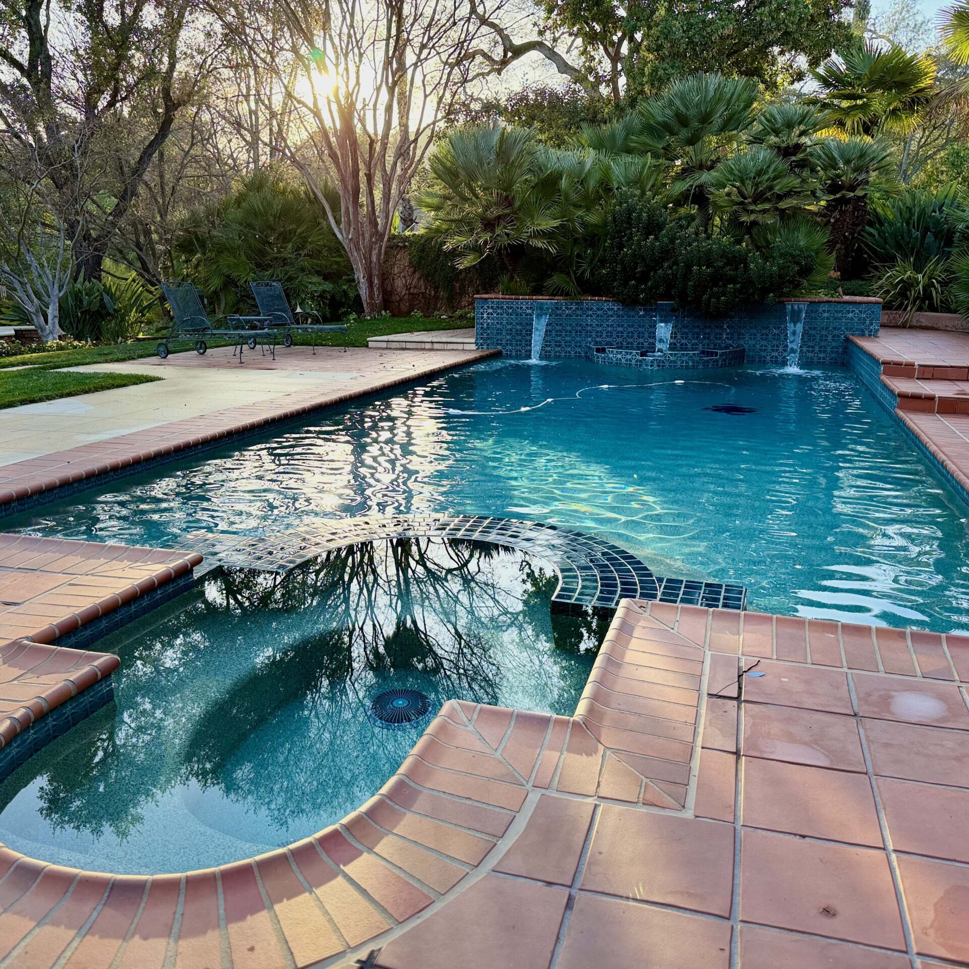 A serene backyard pool with terracotta tiles, surrounded by lush greenery. Sunlight filters through trees, casting reflections on the water's surface.