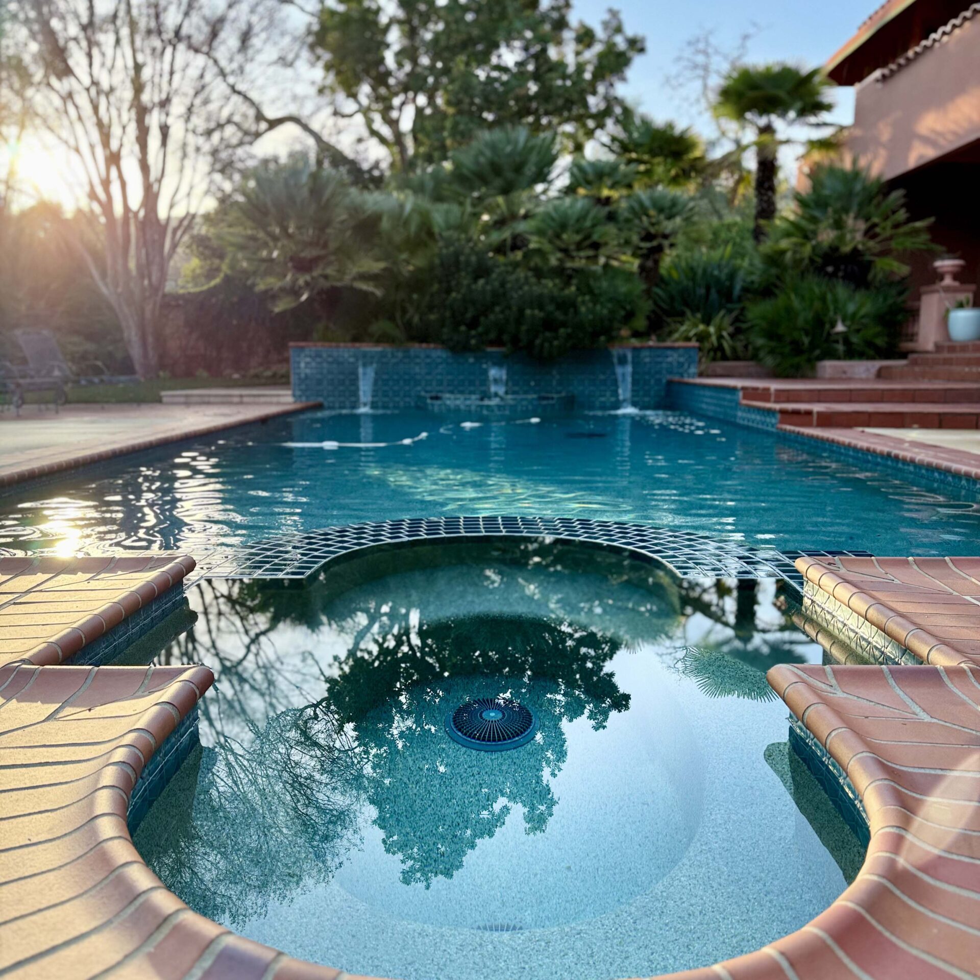 Sunlit swimming pool with a spa section, surrounded by lush greenery and patio, creating a tranquil backyard retreat. No people are present.