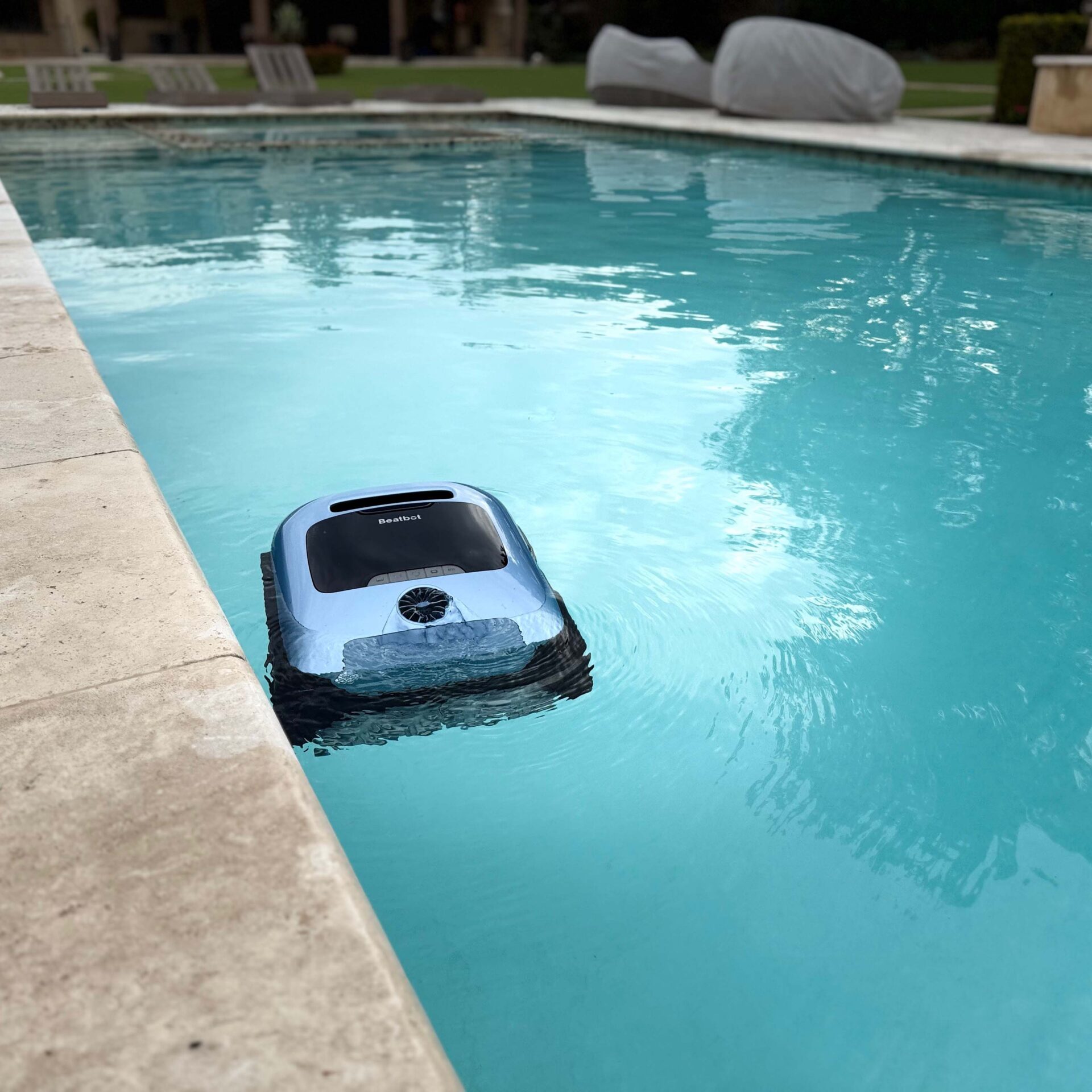 A pool cleaner operates in a backyard swimming pool with clear water and a stone edge, surrounded by lawn chairs and greenery.