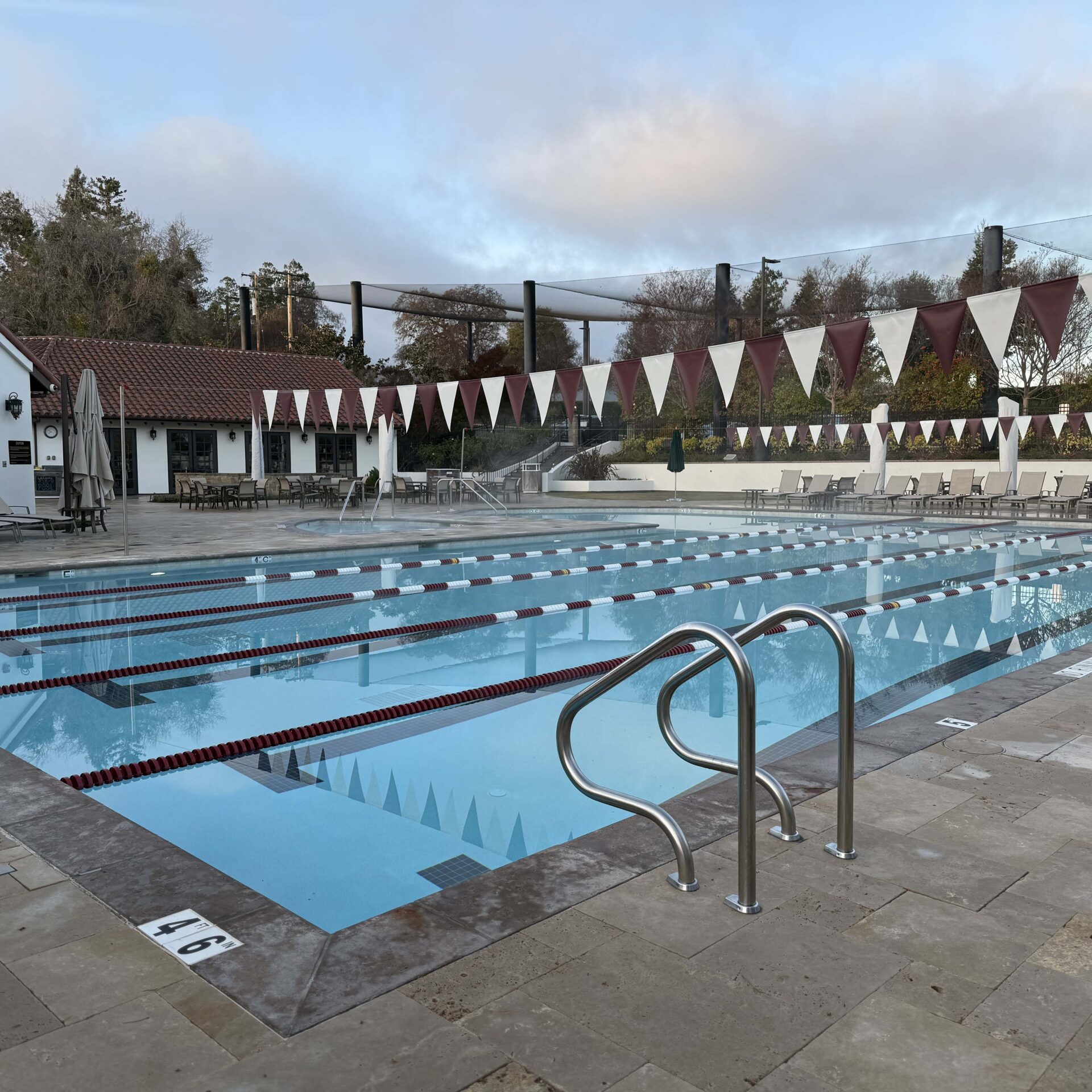 Outdoor swimming pool with lane dividers, surrounded by trees and a building. Lounge chairs line the poolside. Overcast sky overhead.